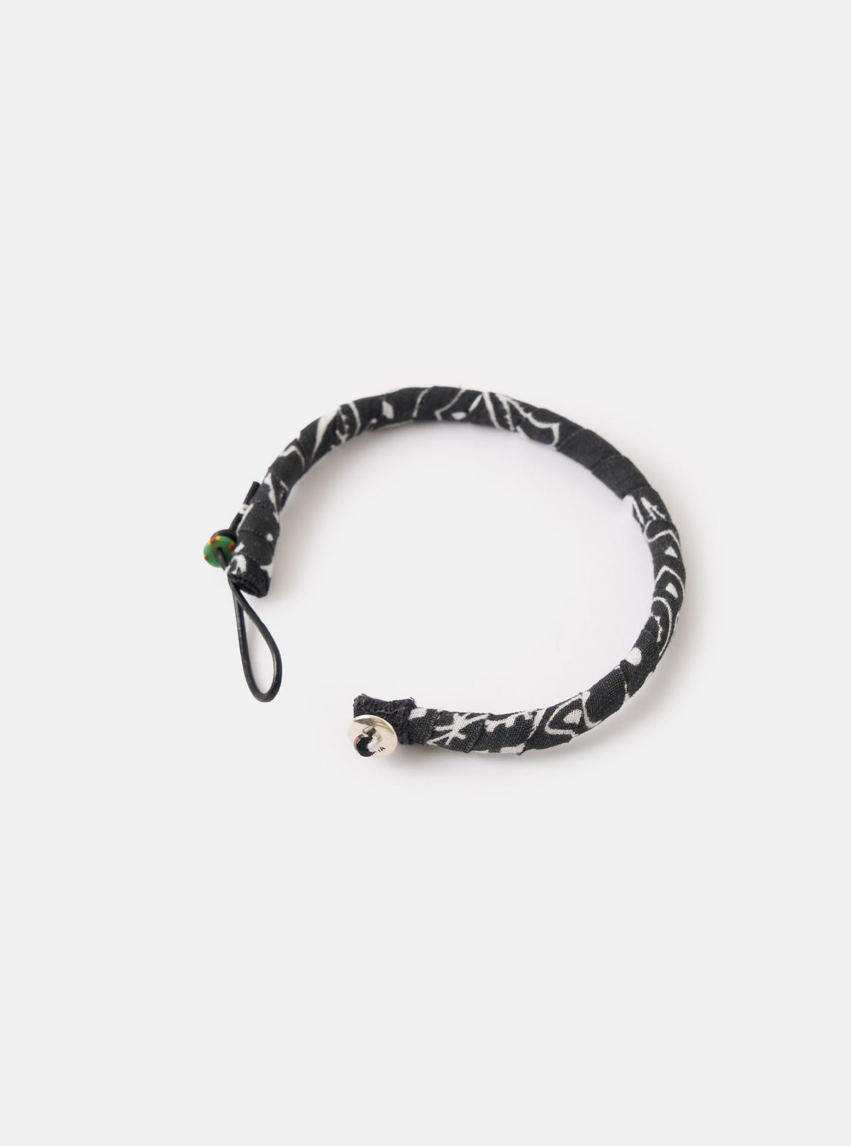 The Mikia Black Bandana Bracelet on a white surface. The bracelet is black with a white paisley pattern and has a silver button closure and black string.