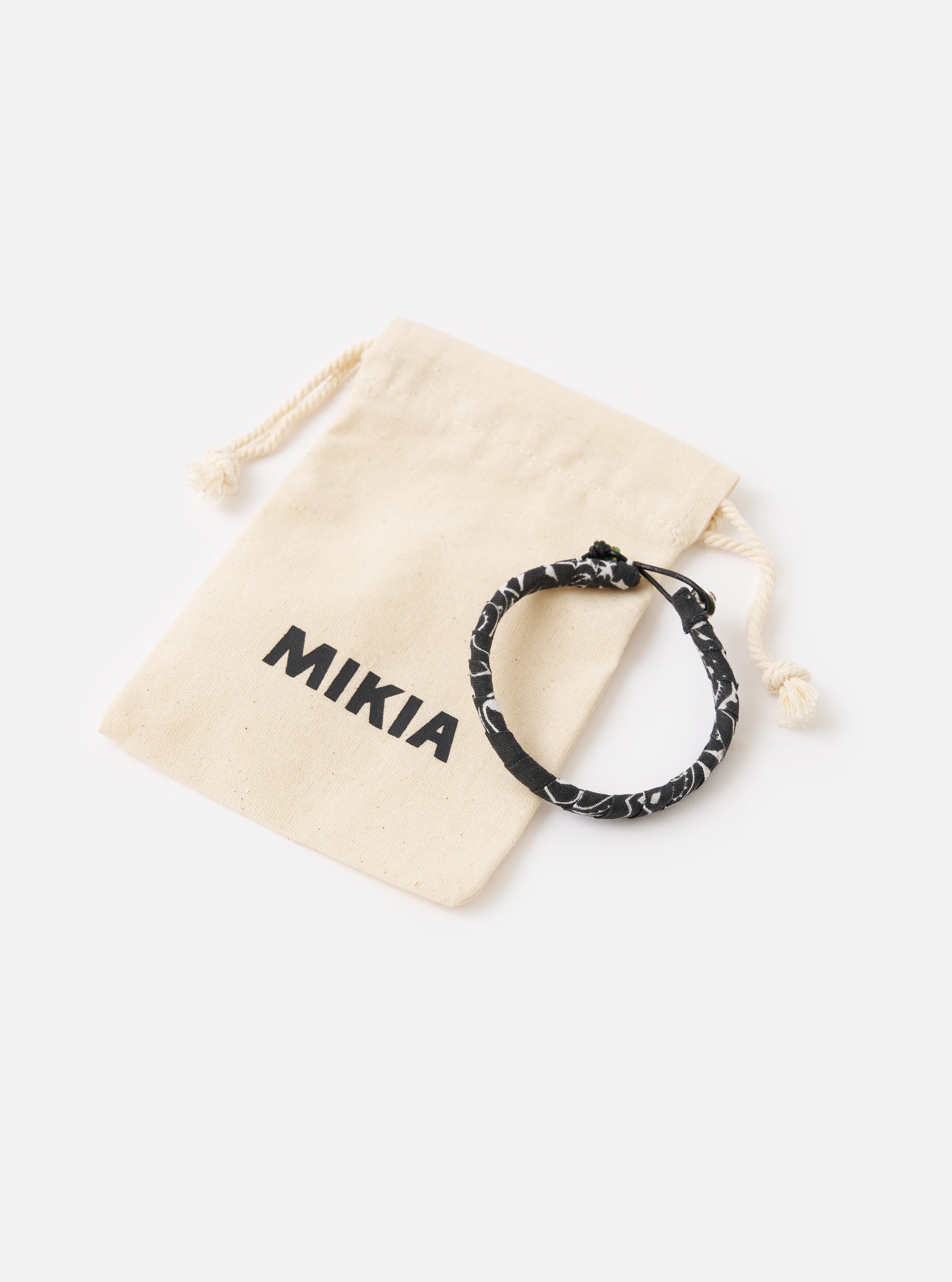 The Mikia Black Bandana Bracelet sits next to its branded dust bag on a white surface. The bracelet is black with a white pattern.