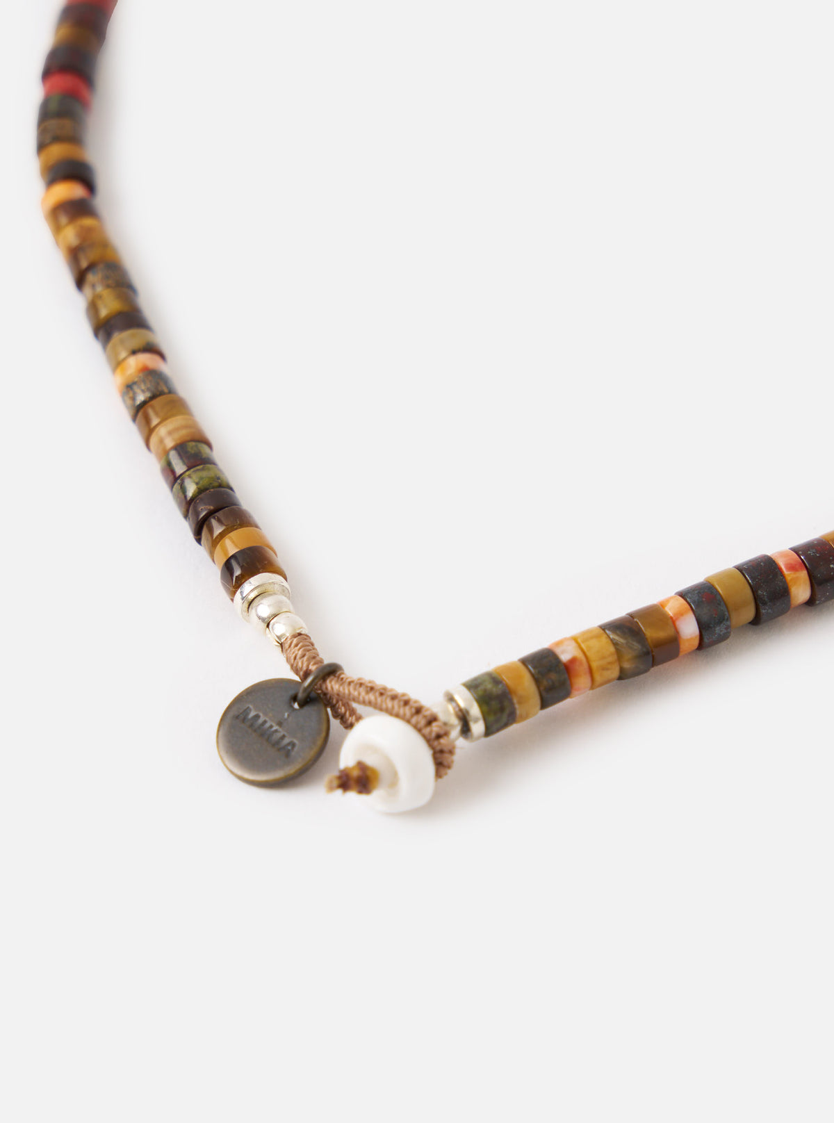 Close-up of the Mikia Brown Multi Heishi Beads Necklace on a white surface. The necklace features brown, red, and orange beads, with a shell and Mikia tag near the clasp.
