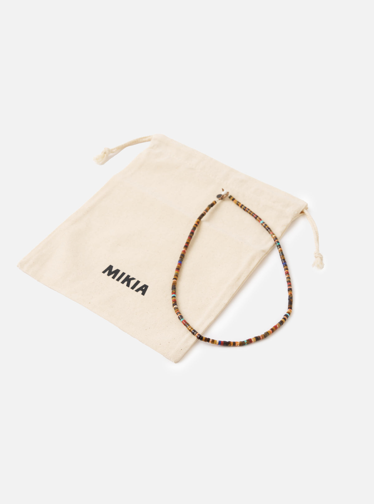 The Mikia Brown Multi Heishi Beads Necklace on a white surface next to its branded beige drawstring bag. The necklace features brown, red, and blue beads.