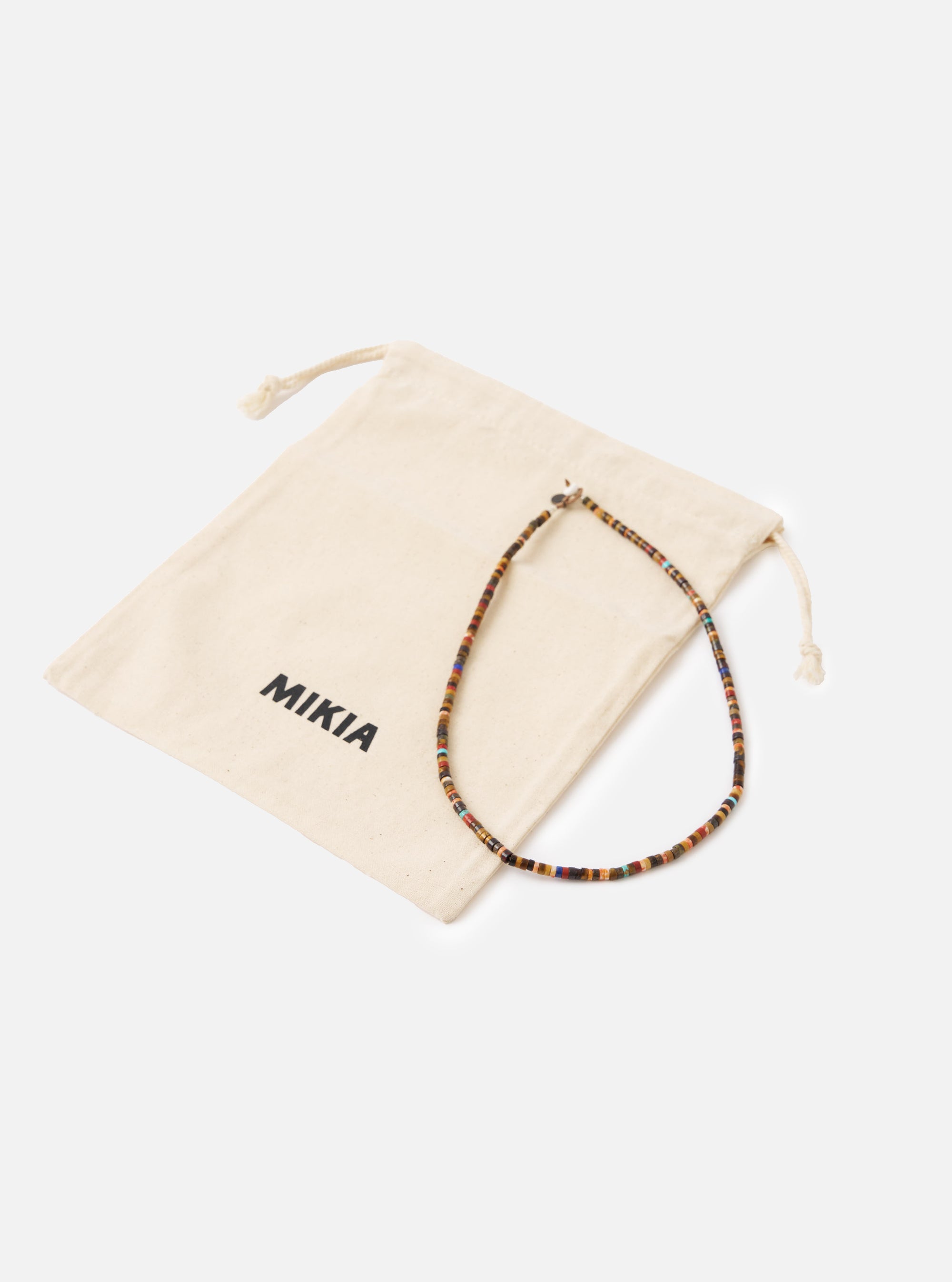 The Mikia Brown Multi Heishi Beads Necklace on a white surface next to its branded beige drawstring bag. The necklace features brown, red, and blue beads.