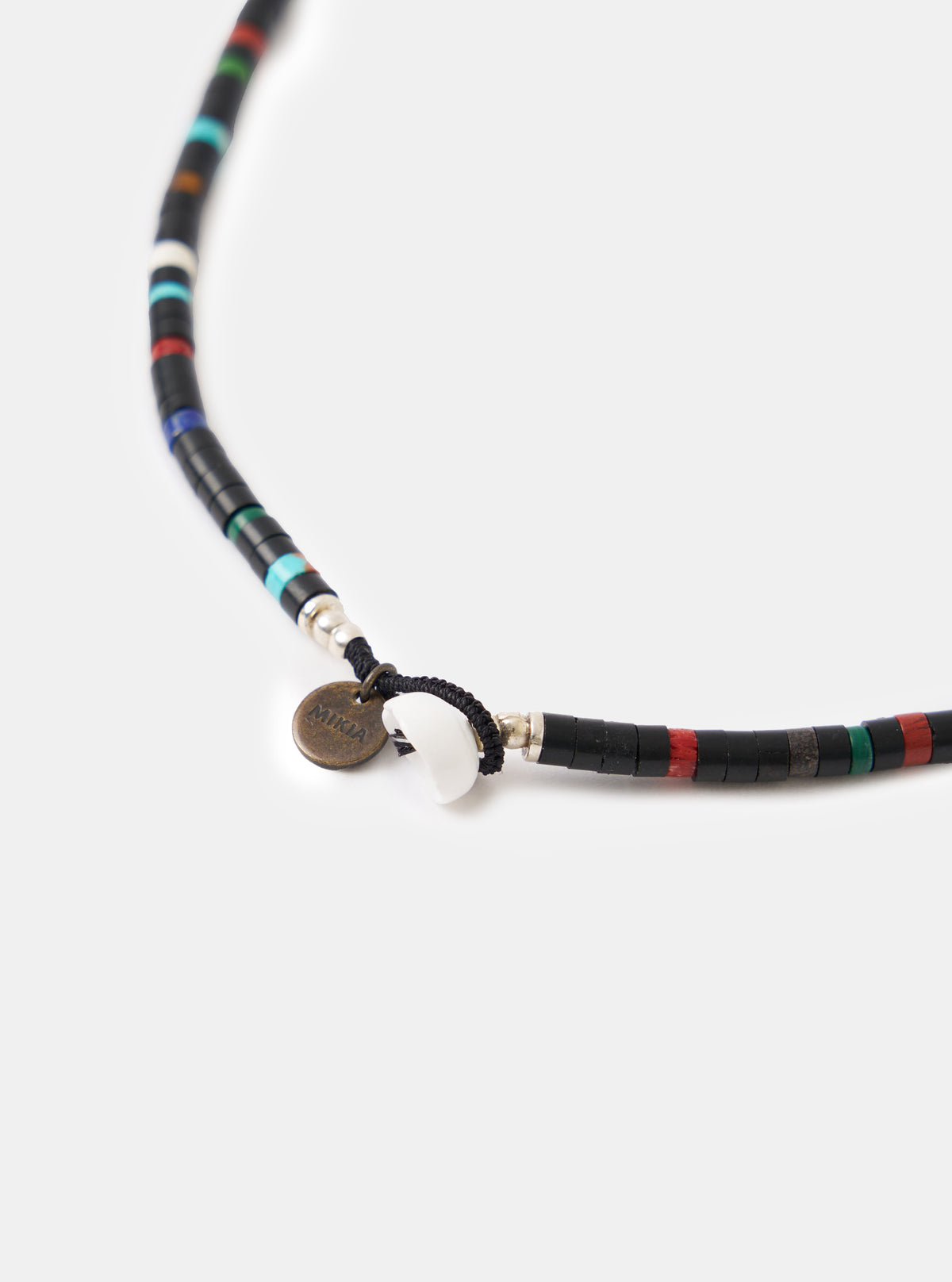 Close-up of the Mikia Jet Multi Heishi Beads Necklace on a white surface. Features black, red, green, and turquoise beads, a white shell, and a Mikia logo tag.