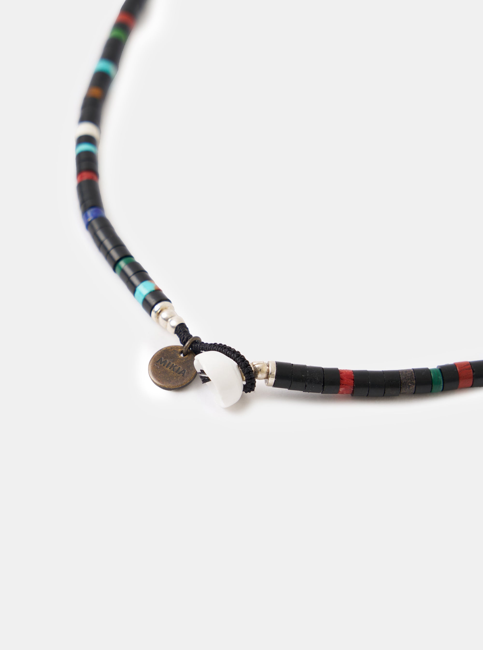 Close-up of the Mikia Jet Multi Heishi Beads Necklace on a white surface. Features black, red, green, and turquoise beads, a white shell, and a Mikia logo tag.