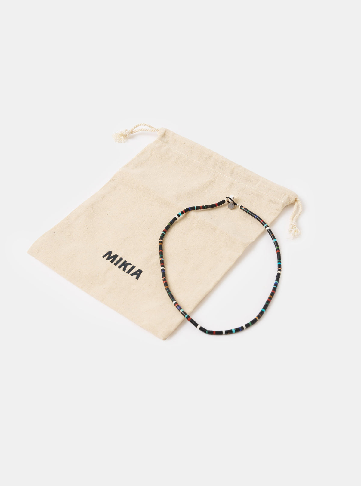The Mikia Jet Multi Heishi Beads Necklace on a white surface next to its branded beige pouch. The necklace features black beads with accents of red, white, and blue.