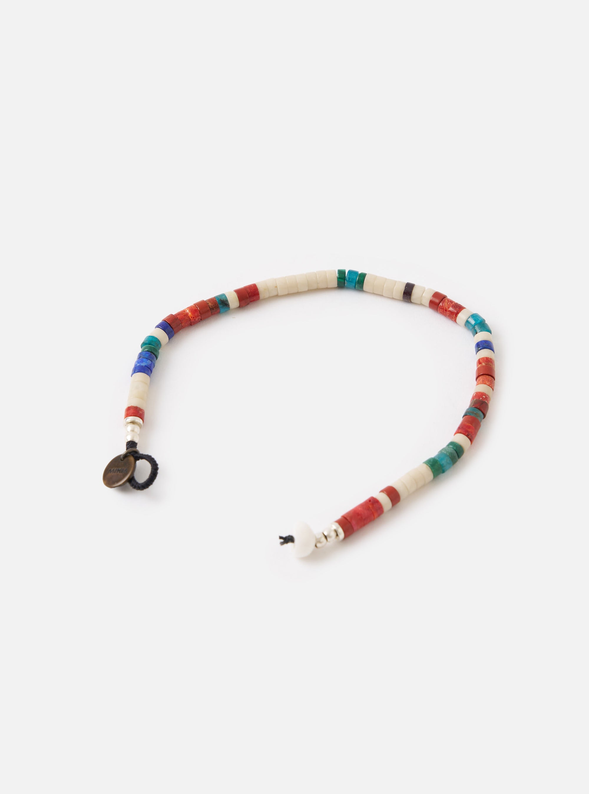 The Mikia Safari Heishi Beads Bracelet on a white surface. The bracelet features colorful heishi beads in red, blue, green, and white. It has a bronze Mikia logo charm.