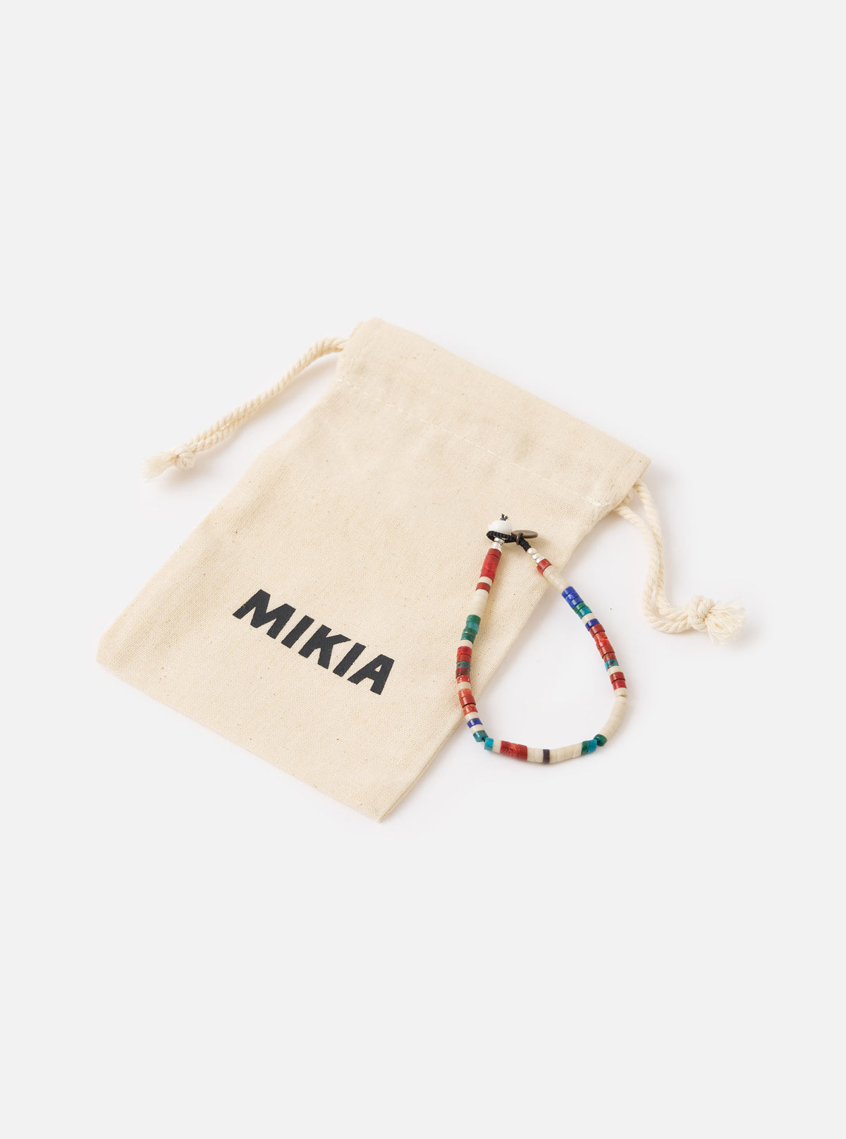 The Mikia Safari Heishi Beads Bracelet on a white surface next to its branded beige pouch. The bracelet features red, white, blue and green beads.