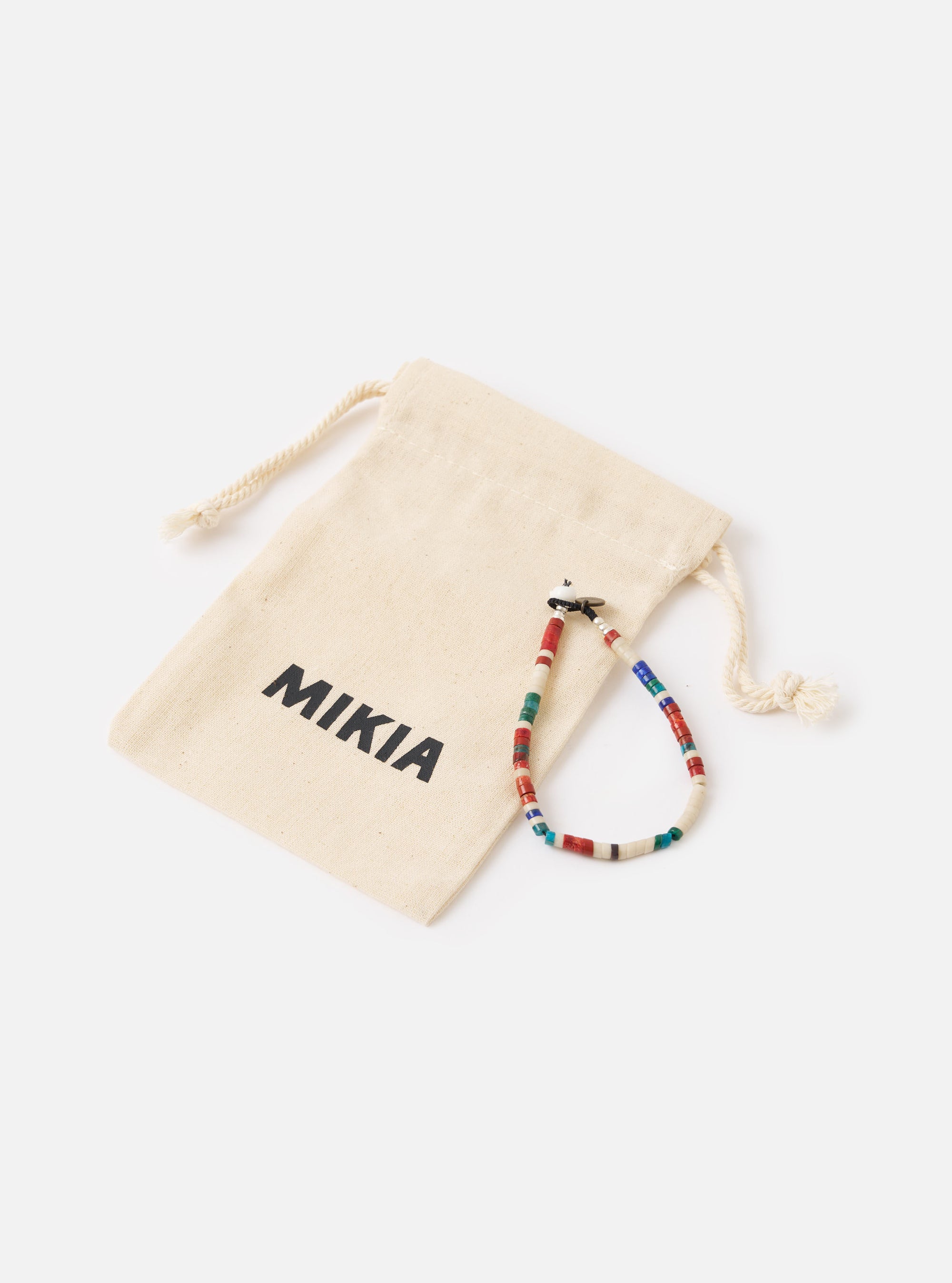 The Mikia Safari Heishi Beads Bracelet on a white surface next to its branded beige pouch. The bracelet features red, white, blue and green beads.