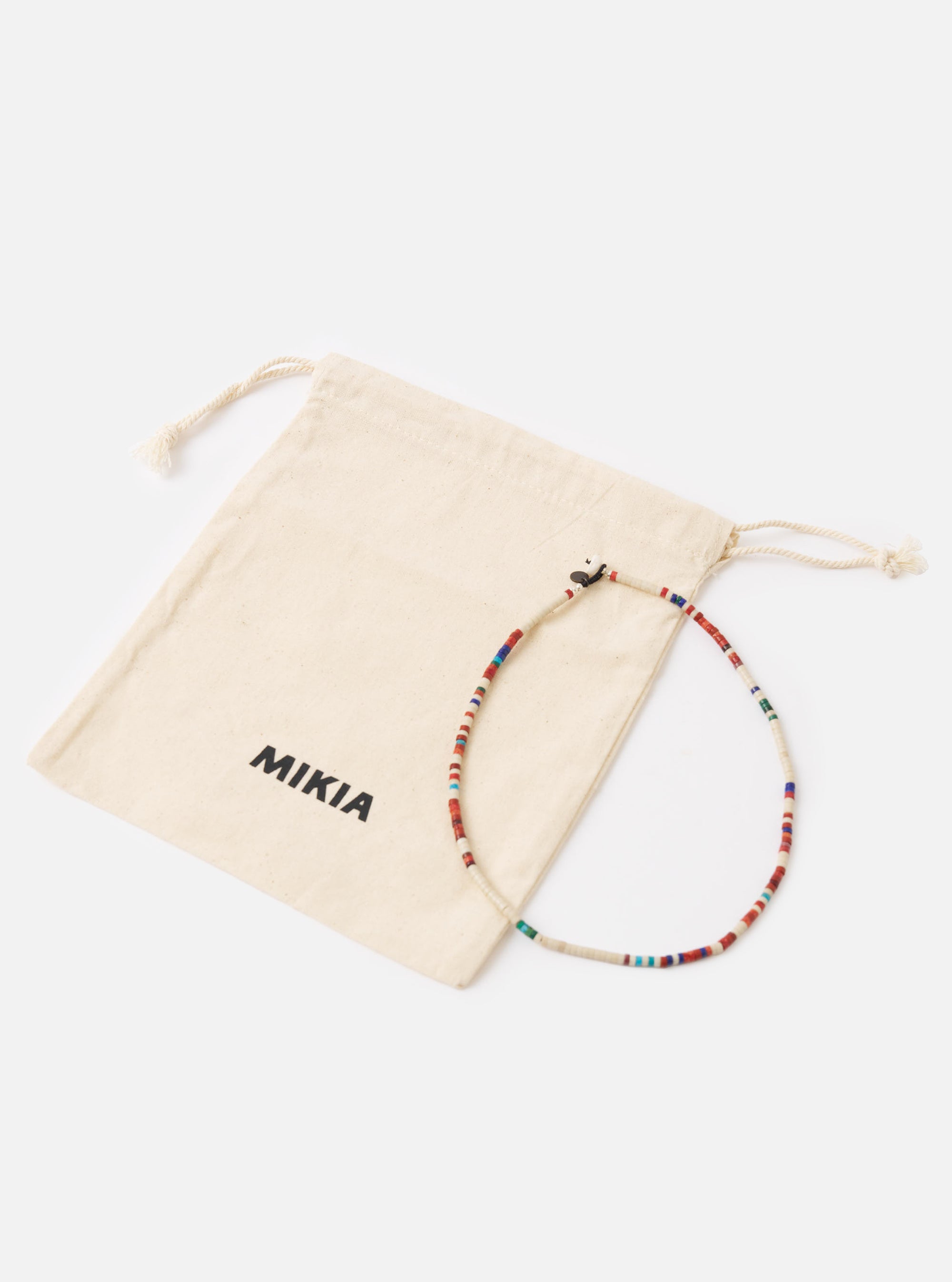 The Mikia Safari Heishi Beads Necklace with red, white and blue beads, next to its branded beige pouch on a white surface.