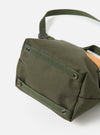 Close-up of the base of a master-piece x Universal Works Olive Recycled Tech Canvas Small Tote Bag on a white surface. The olive green canvas is detailed with webbing and metal rivets.