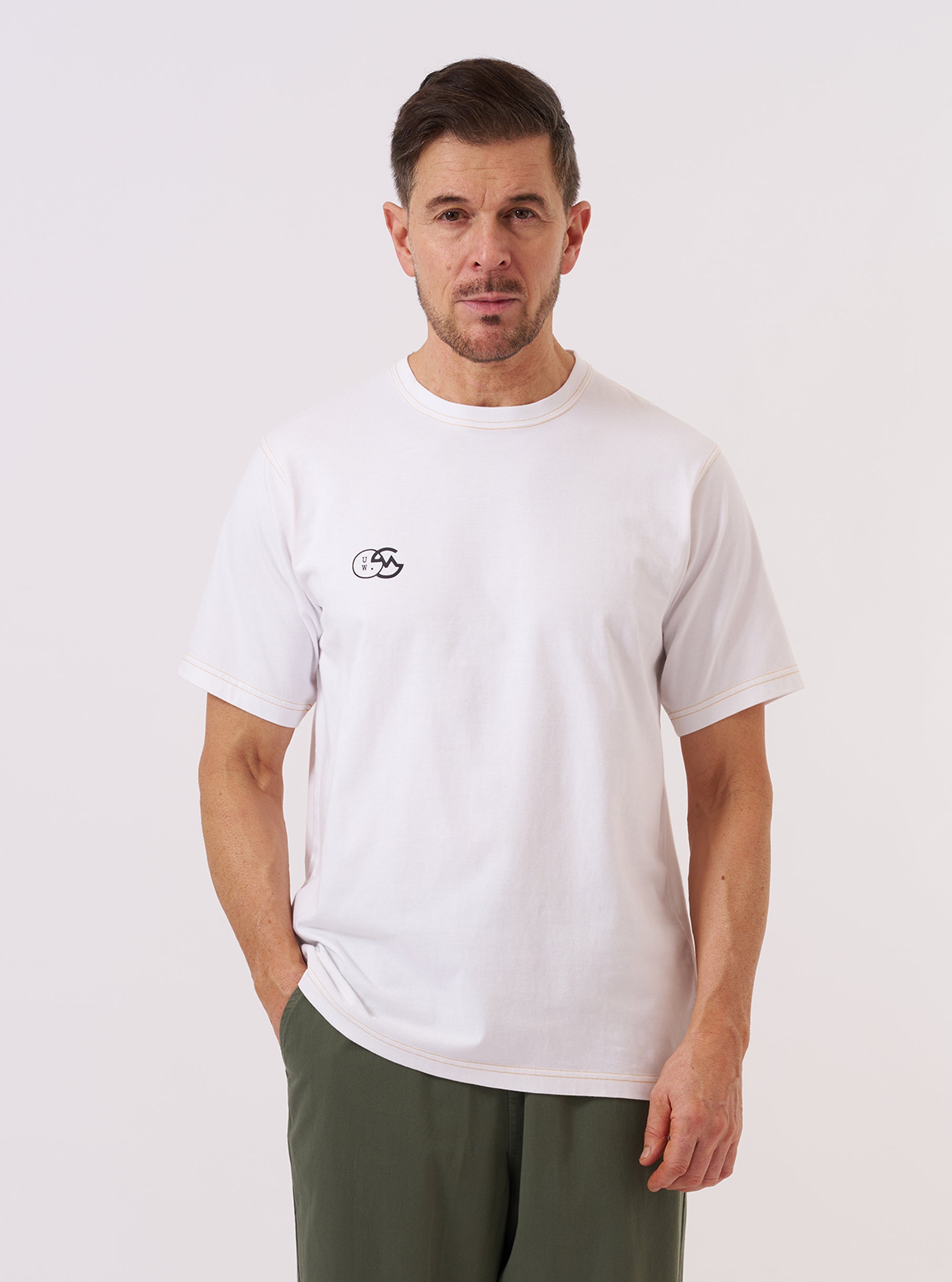 A model wears the master-piece x Universal Works White Single Jersey Print Tee against a plain white background. The T-shirt is a short-sleeved, crewneck style featuring a large circular graphic on the front with the words Master-piece and Universal Works encircling a central design. The model is shown from the waist up, and the garment has a relaxed fit. No mannequin is present in the image.