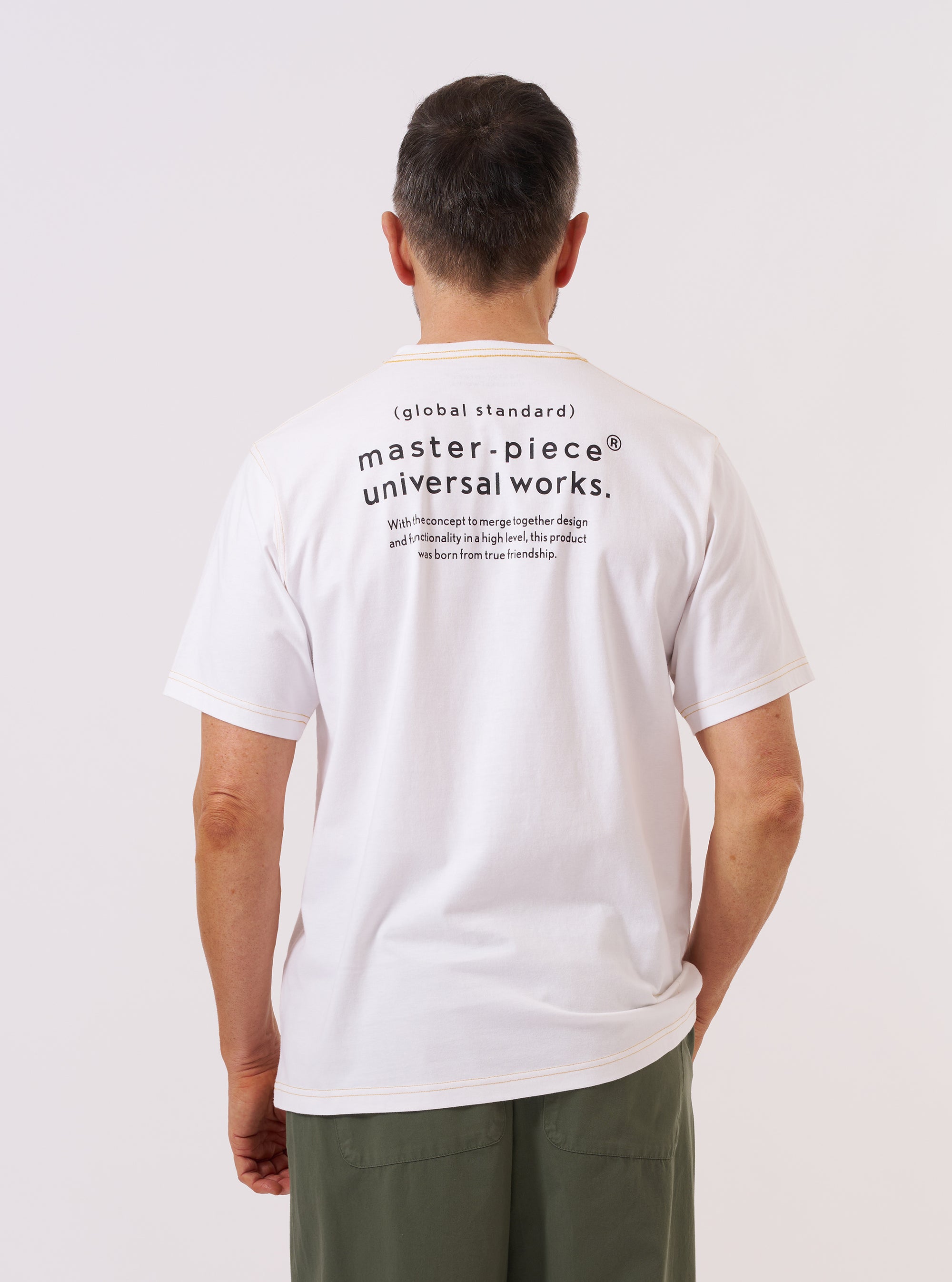 A model wears the master-piece x Universal Works White Single Jersey Print Tee, shown from the chest down against a plain white background. The white short-sleeve T-shirt features a large graphic print on the front with "UNIVERSAL WORKS" text above a circular design and "MASTER-PIECE" text below. The model is also wearing dark olive trousers.