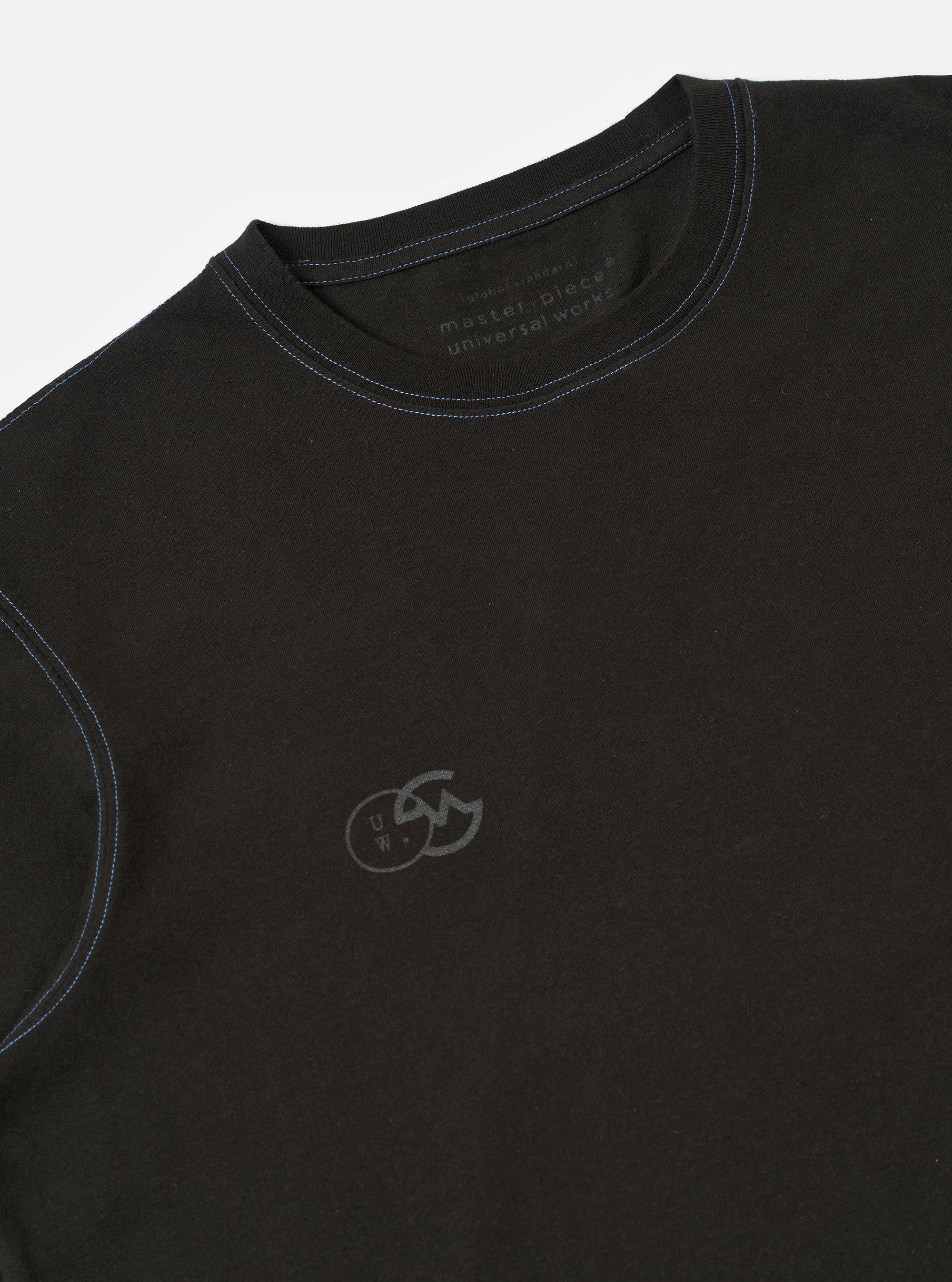 The master-piece x Universal Works Black Single Jersey Print Tee is shown as a flat lay against a white background. The image displays the back of the short-sleeved t-shirt, highlighting a small, circular dual-branded logo printed in white near the neckline and a white woven master-piece tag stitched at the bottom hem.