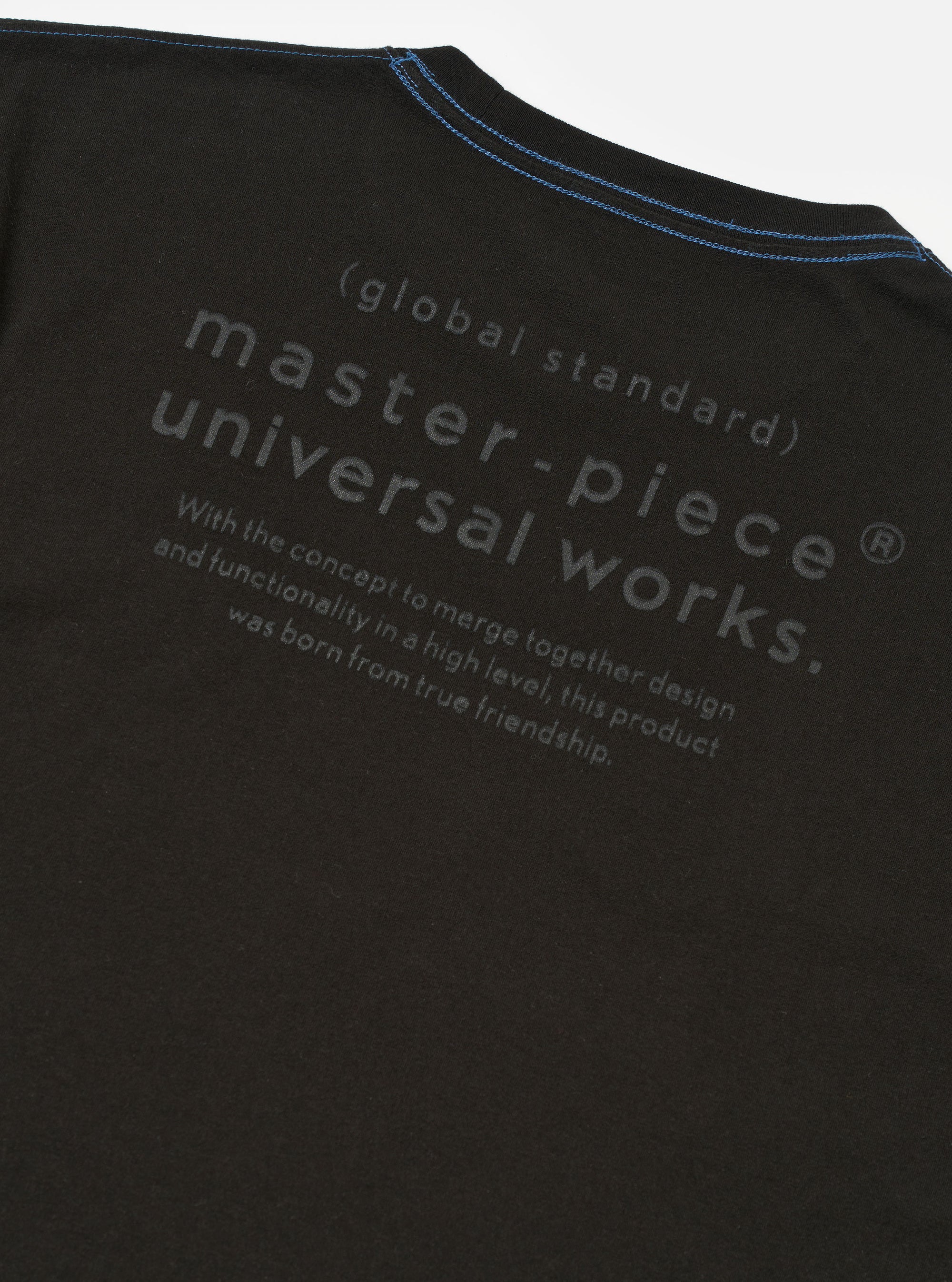 The master-piece x Universal Works Black Single Jersey Print Tee is shown from the back as a flat lay against a plain white background. The image features a large graphic print centered on the upper back, which includes the word master-piece in a stylized font above smaller text and a collaboration logo. The t-shirt is a short-sleeved crew neck made from black cotton fabric with a smooth finish.