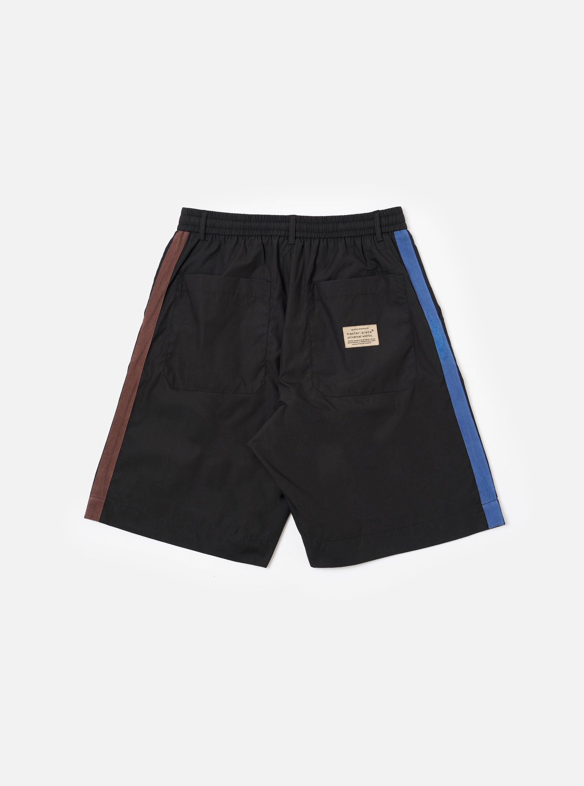 The master-piece x Universal Works Black Recycled Poly Tech Pleated Track Short is shown from the back as a flat lay against a white background. The image highlights the elasticated waistband and two rear pockets, including one with a button-down flap. The black recycled poly-tech fabric has a smooth, utilitarian finish.