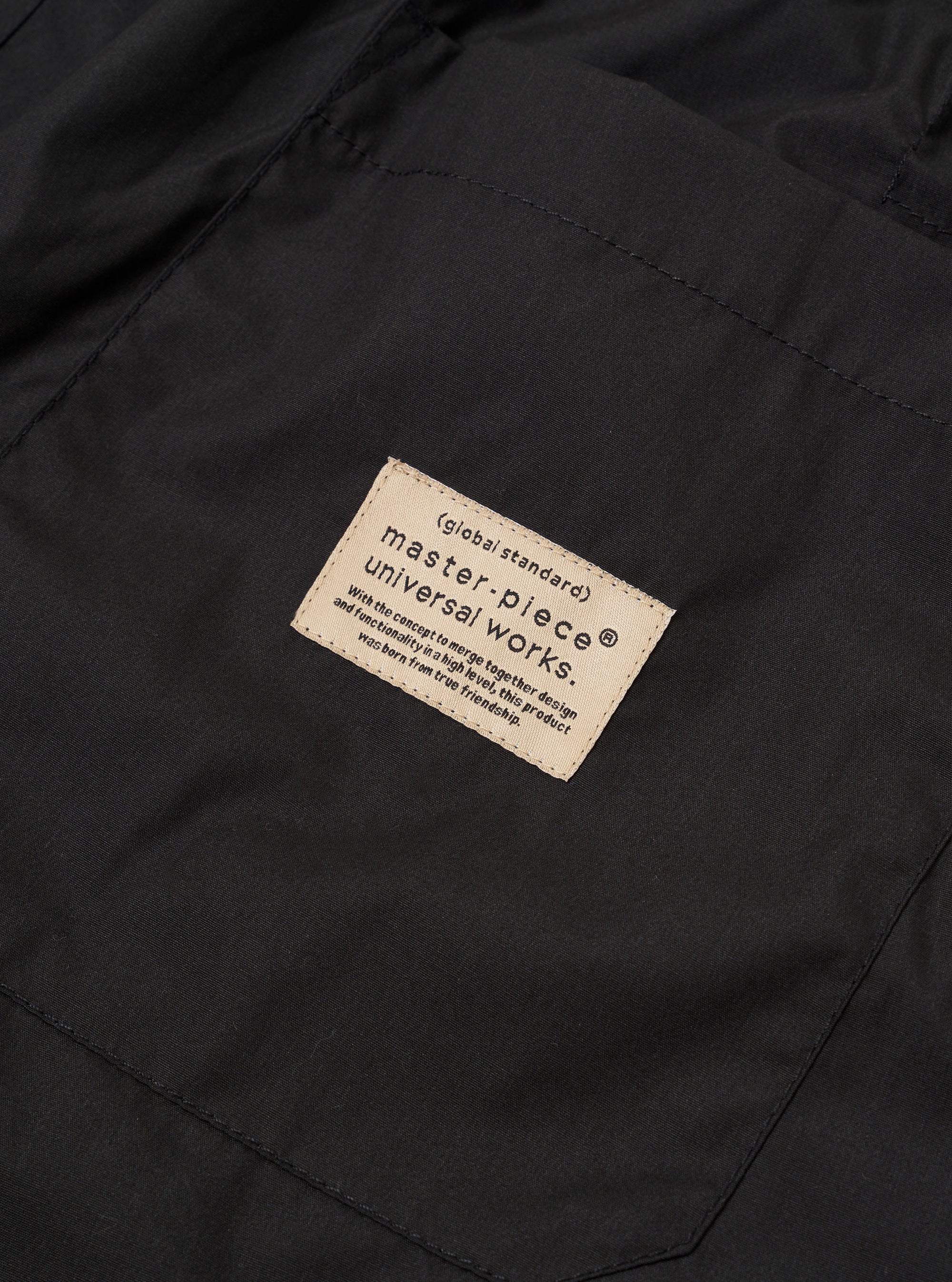 The master-piece x Universal Works Black Recycled Poly Tech Pleated Track Short is shown from a rear angle as a flat lay against a white background. The image highlights the two rear pockets, the elasticated waistband with belt loops, and the loose, relaxed fit of the black fabric.
