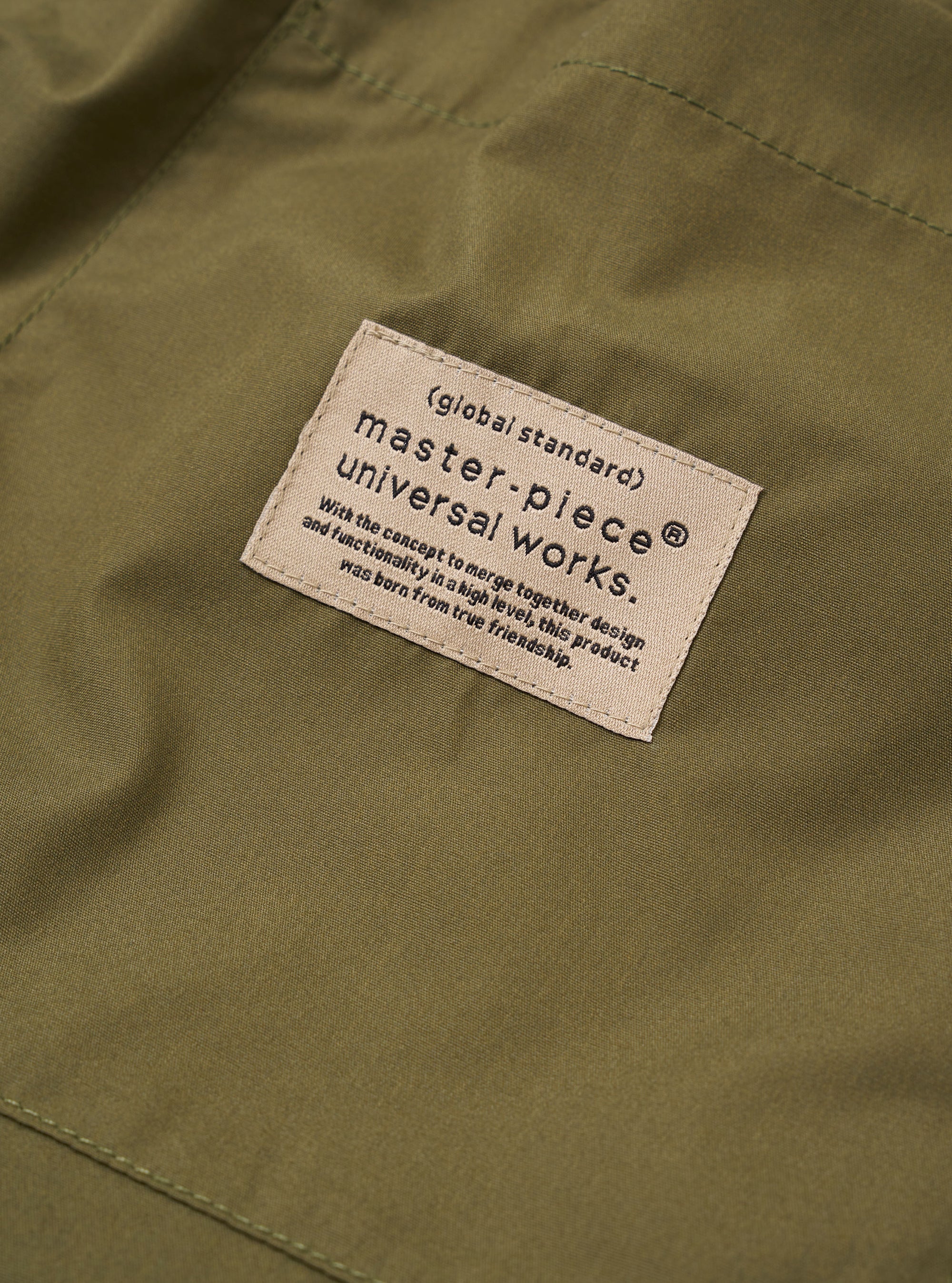 The master-piece x Universal Works Olive Recycled Poly Tech Pleated Track Short is shown from the back as a flat lay against a white background. The image highlights the two rear pockets, one of which has a button closure, and the elasticated waistband with belt loops.