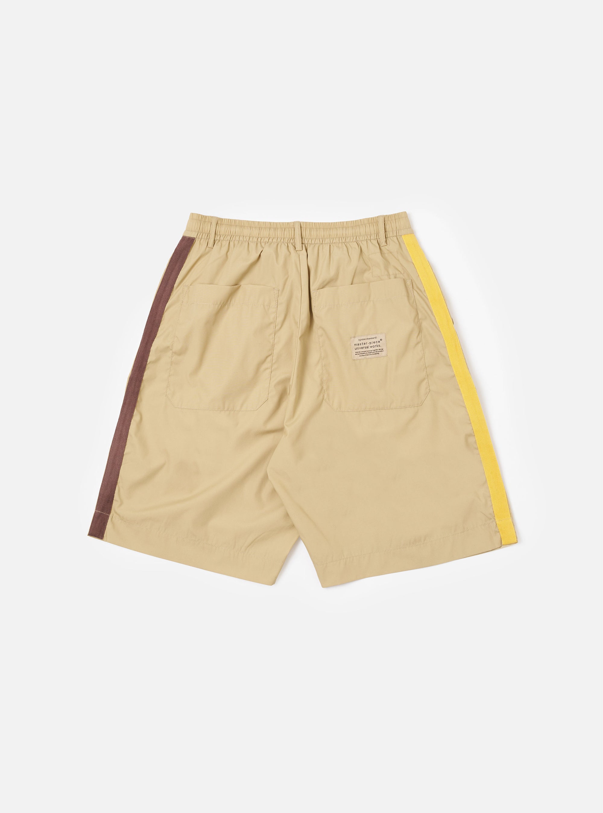 The master-piece x Universal Works Sand Recycled Poly Tech Pleated Track Short is shown from the back as a flat lay against a white background. The image highlights the two rear pockets, including one with a button-down flap, and the elasticated waistband. The sand-colored fabric has a smooth, utilitarian finish.