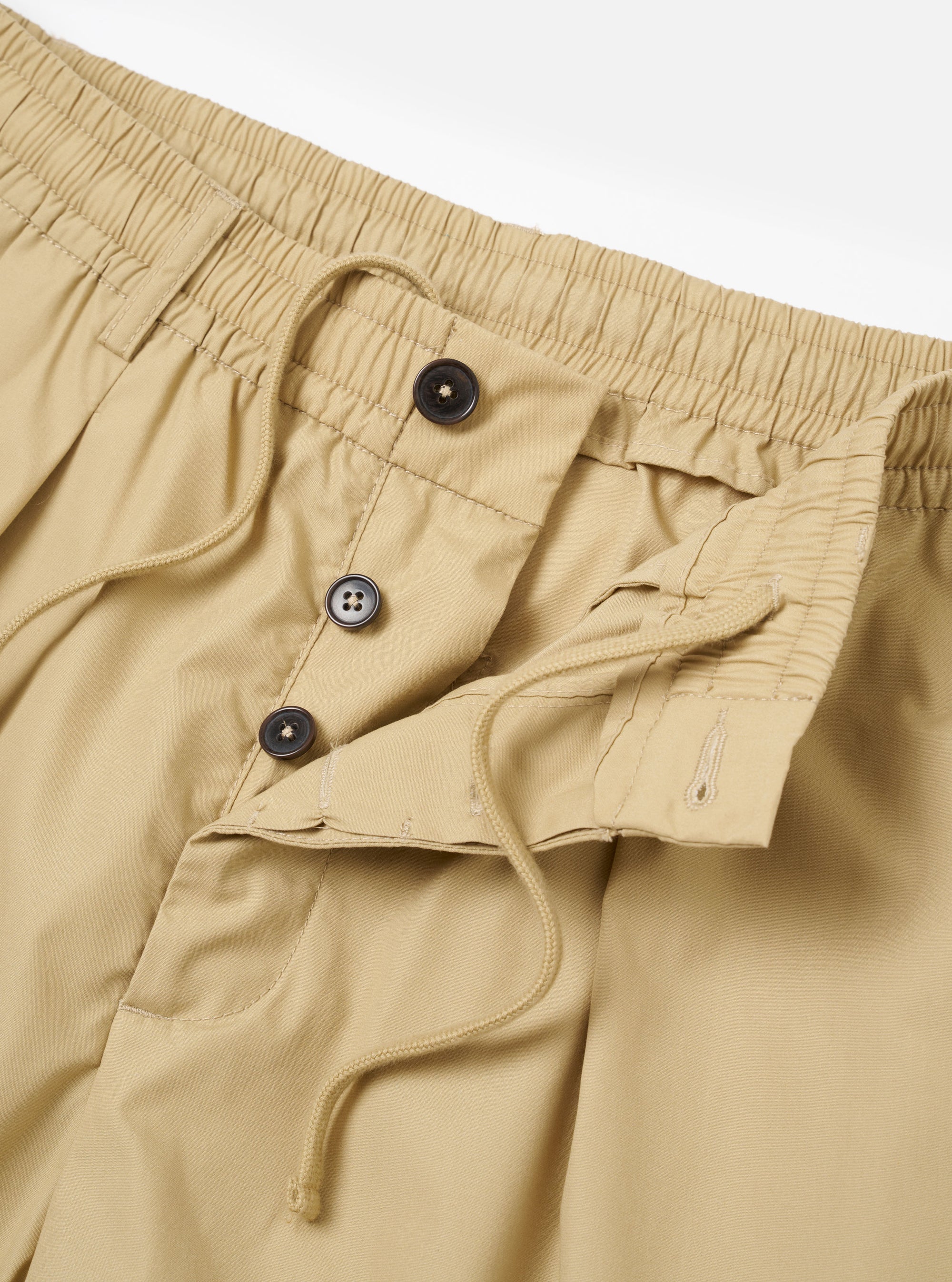 The master-piece x Universal Works Sand Recycled Poly Tech Pleated Track Short is shown from a rear three-quarter angle as a flat lay against a plain white background. The image highlights the relaxed fit, the elasticated waistband with a white drawstring, and a single rear welt pocket.