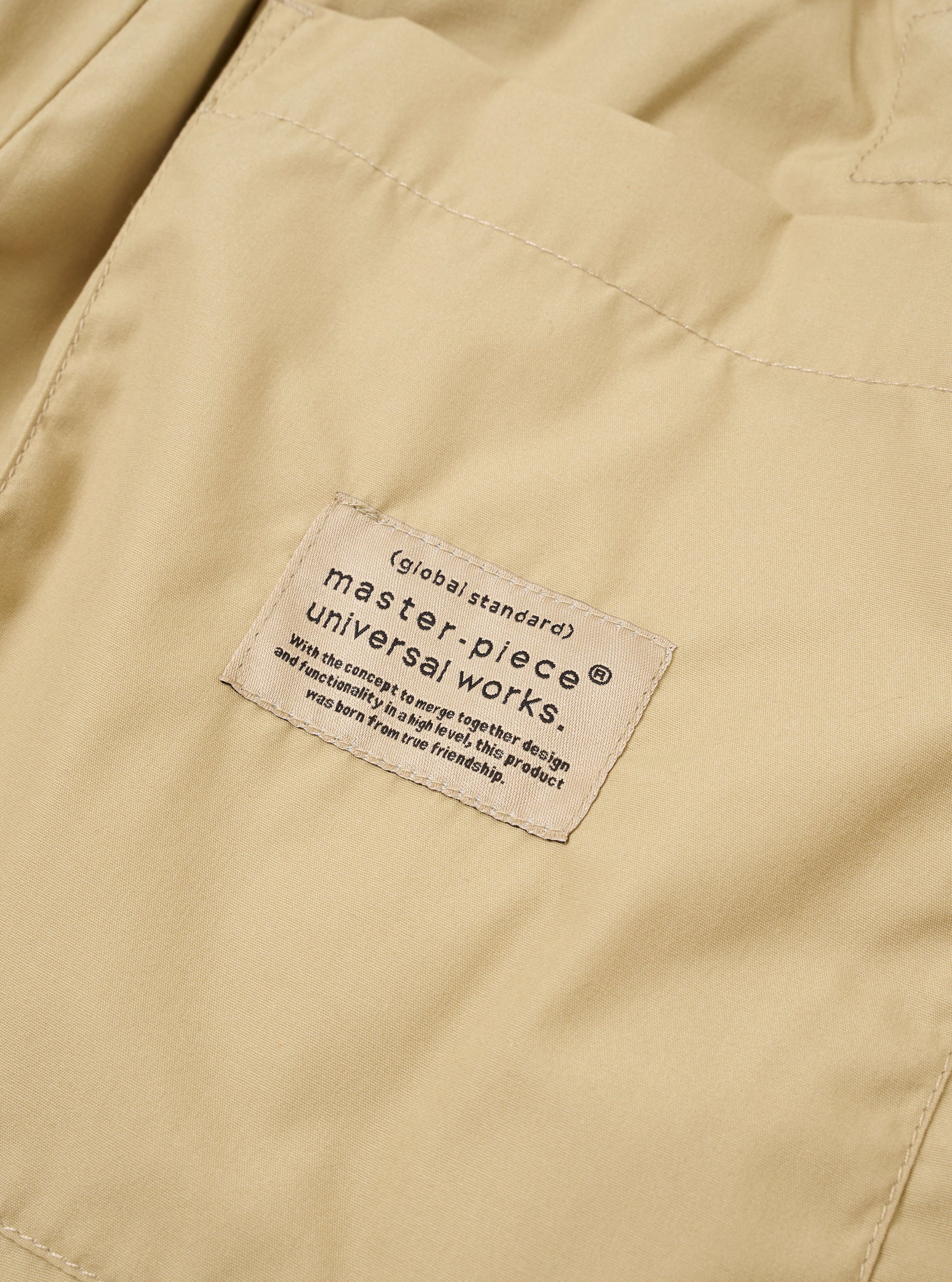 The master-piece x Universal Works Sand Recycled Poly Tech Pleated Track Short is shown from the back as a flat lay against a white background. The image highlights the two rear pockets, including one with a button-down flap, and the elasticated waistband.