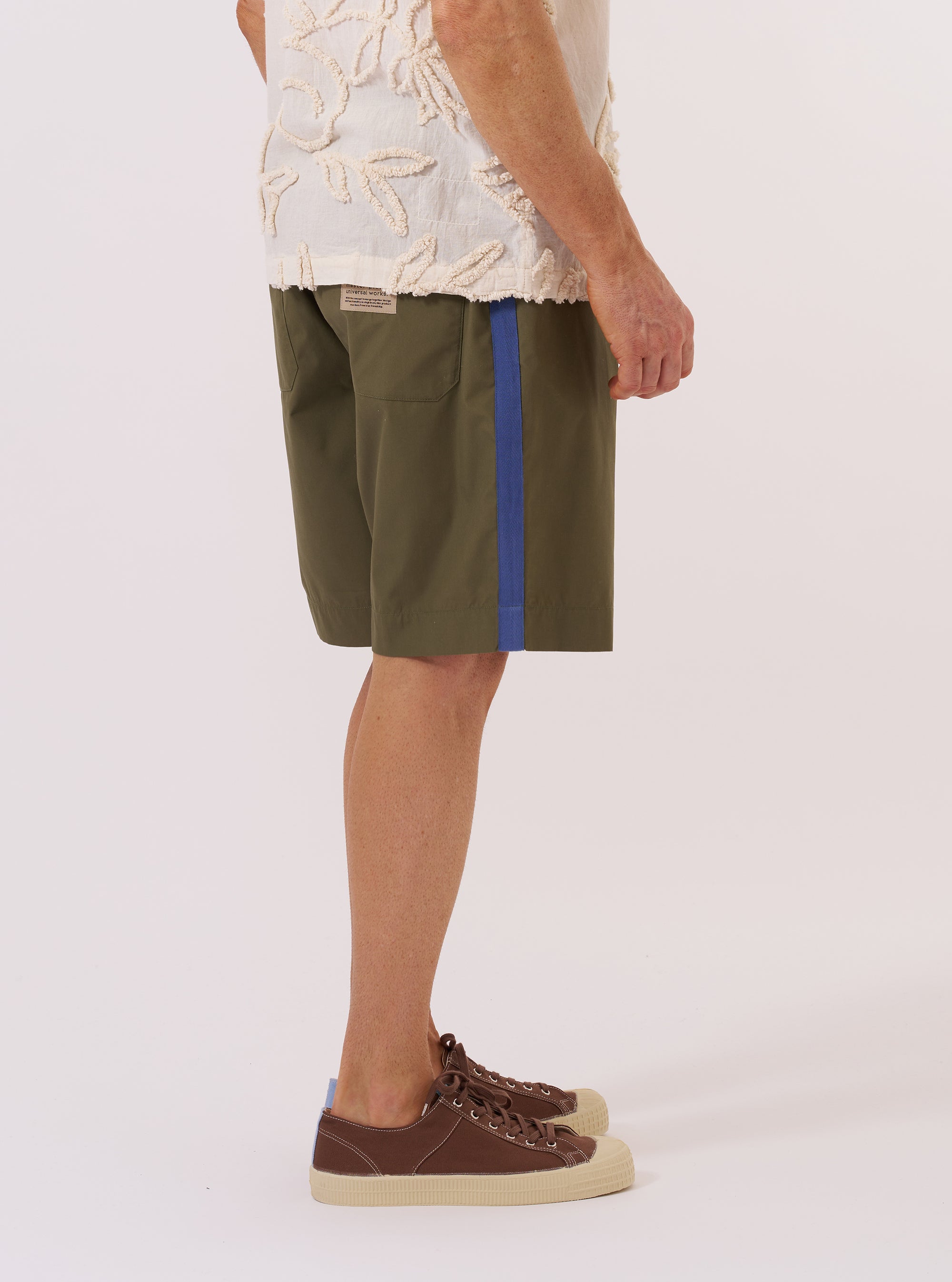 A model wears the master-piece x Universal Works Olive Recycled Poly Tech Pleated Track Short, shown from the waist down against a plain white background. The olive green shorts feature a relaxed fit, a single front pleat on each leg, and an elasticated waistband with a button closure. The model is wearing the shorts with white socks and a white T-shirt tucked in to highlight the waistband and pocket details.