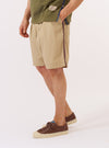 The master-piece x Universal Works Sand Recycled Poly Tech Pleated Track Short is worn by a model against a plain white background. The image shows the model from the waist down, highlighting the shorts' relaxed fit, single pleat front, and elasticated drawstring waistband. The sand-colored fabric has a smooth, technical finish. Front slant pockets are visible at the sides.