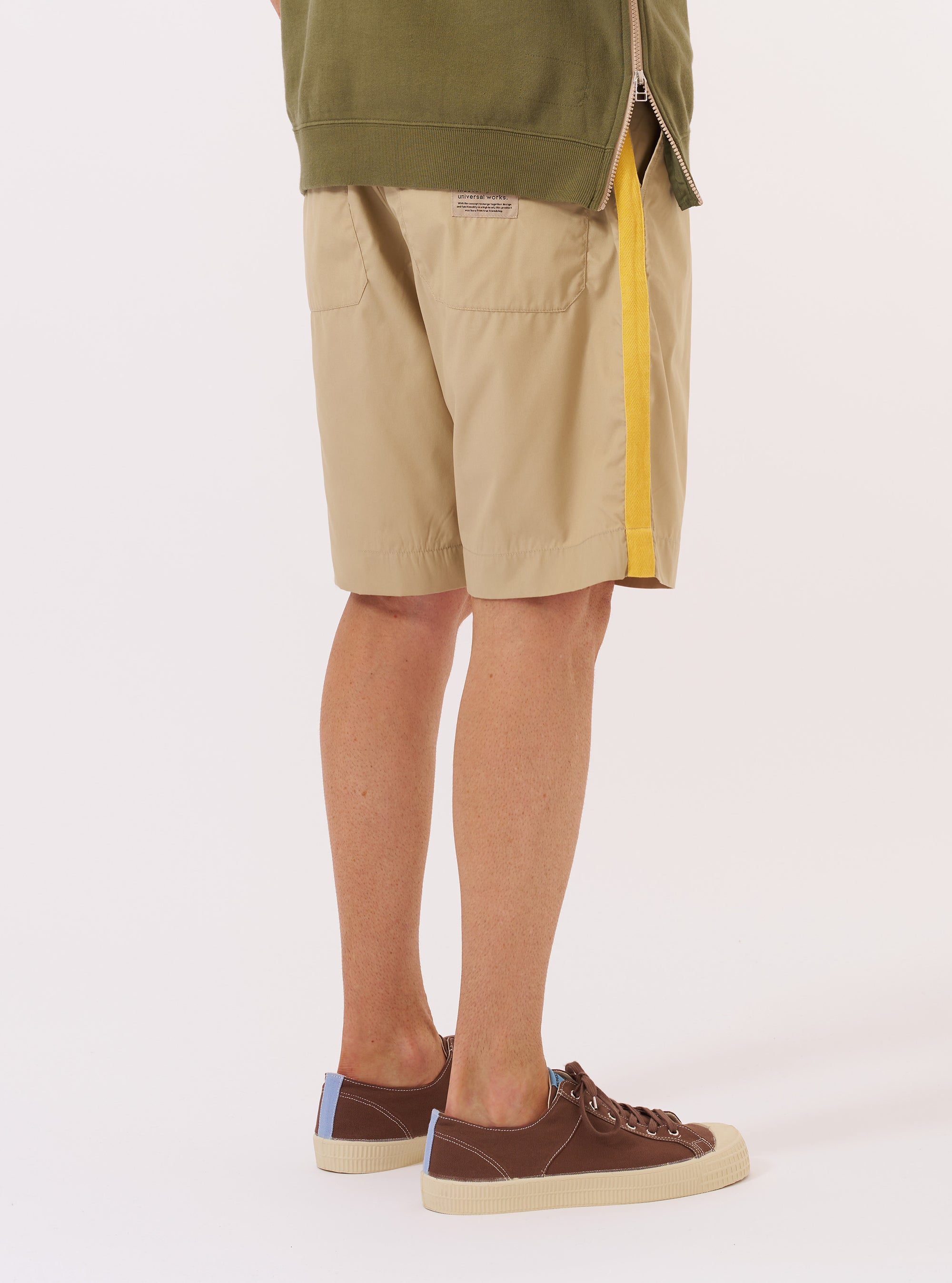 The master-piece x Universal Works Sand Recycled Poly Tech Pleated Track Short is worn by a model against a plain white background. The image shows the shorts from the waist down, highlighting the loose, relaxed fit, the pleated front, and the elasticated waistband. The model is also wearing white socks and brown sandals.
