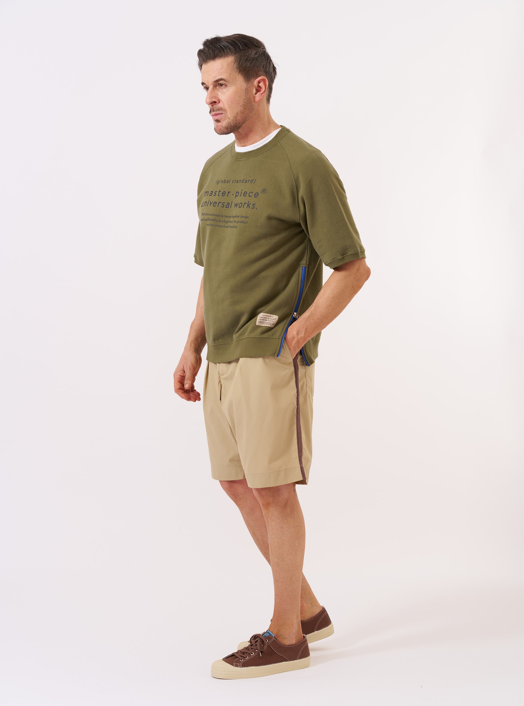 The master-piece x Universal Works Olive Brush Back Sweat S/S Billy Sweat is shown as a flat lay against a white background. This short-sleeved crewneck sweatshirt features a ribbed neckline, a straight hem, and prominent contrasting white zippers at the side seams. The fabric is a textured olive green brush back sweat.