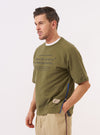 A model wears the master-piece x Universal Works Olive Brush Back Sweat S/S Billy Sweat against a plain white background. The olive green, short-sleeved sweatshirt is worn over a light-colored long-sleeved shirt and features a ribbed crew neck and a chest pocket with a contrasting white logo. The model is shown from the waist up.