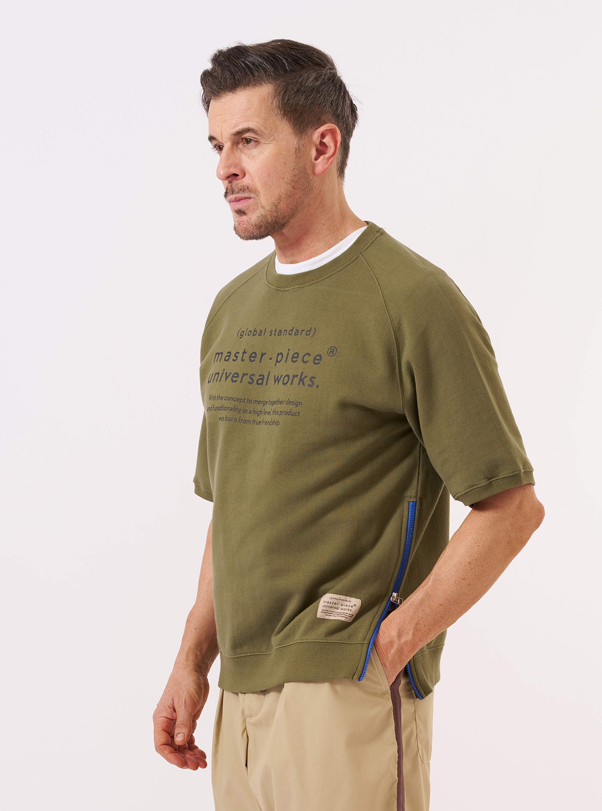 A model wears the master-piece x Universal Works Olive Brush Back Sweat S/S Billy Sweat against a plain white background. The olive green, short-sleeved sweatshirt is worn over a light-colored long-sleeved shirt and features a ribbed crew neck and a chest pocket with a contrasting white logo. The model is shown from the waist up.