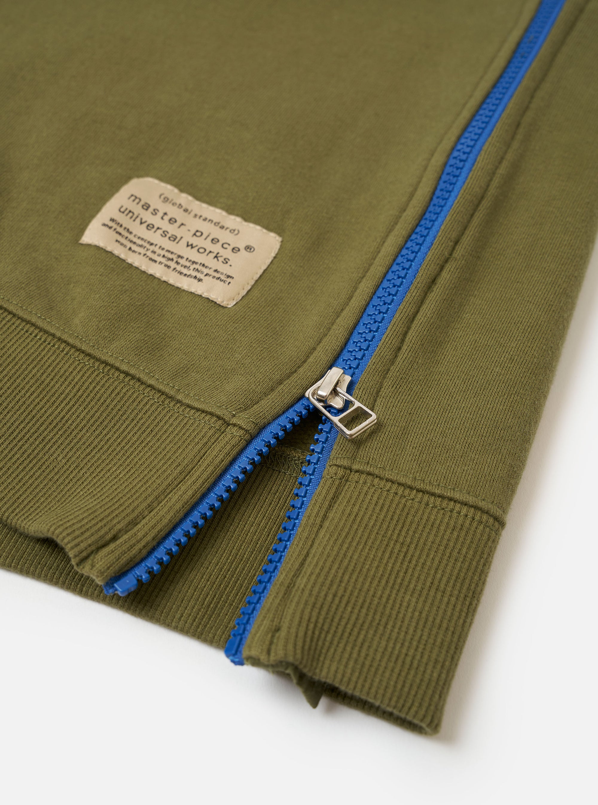 The master-piece x Universal Works Olive Brush Back Sweat S/S Billy Sweat is shown from the back as a flat lay against a plain white background. The short-sleeved sweatshirt features a ribbed crew neck and a straight hem. A small, square collaborative logo patch is positioned on the upper center of the back.