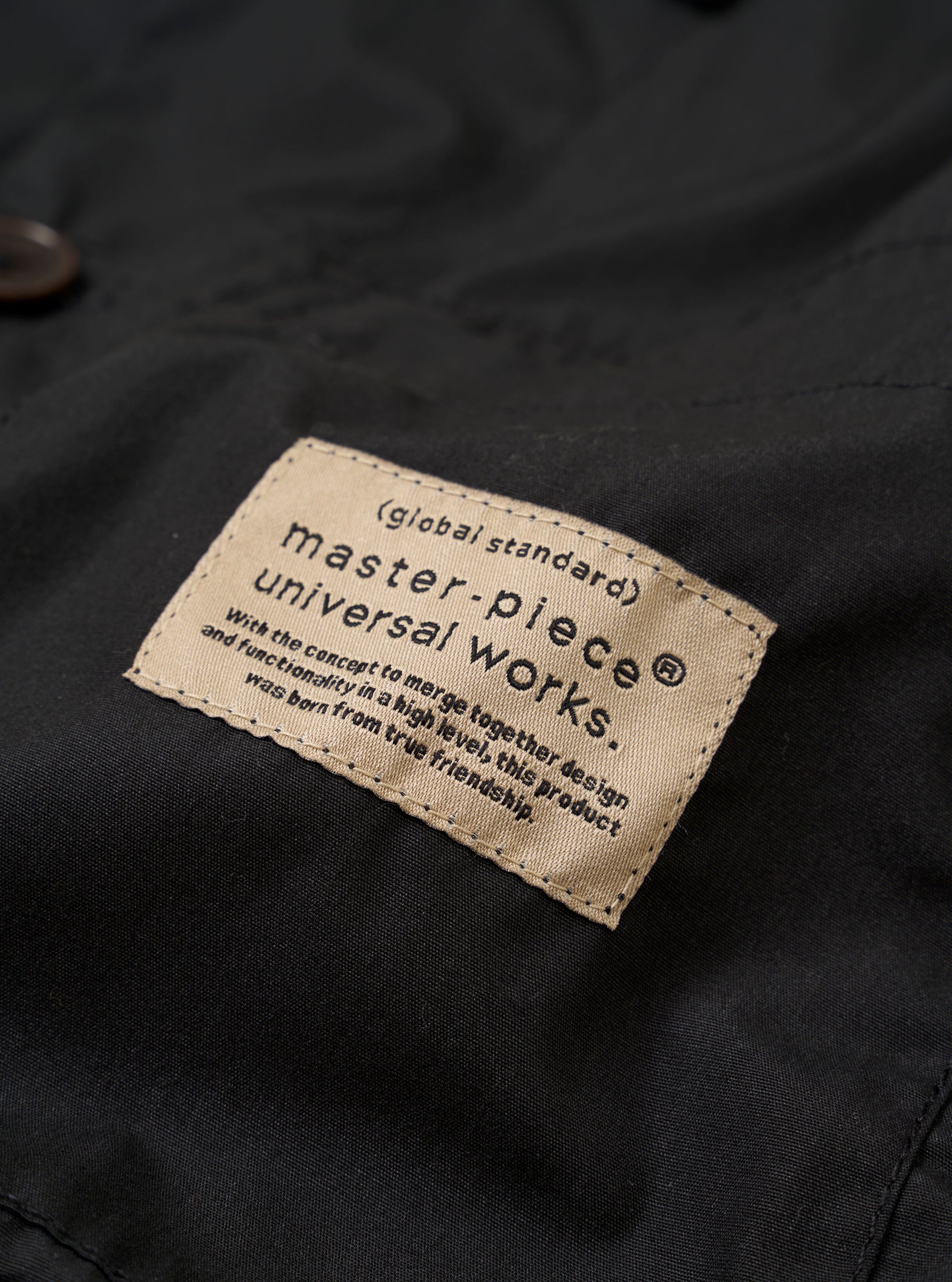The master-piece x Universal Works Black Recycled Poly Tech Tech Overshirt is shown from the back in a studio shot against a plain white background. The image highlights the back yoke with a central box pleat and the elasticated drawcord hem. The overshirt features short sleeves and a structured one-piece collar.