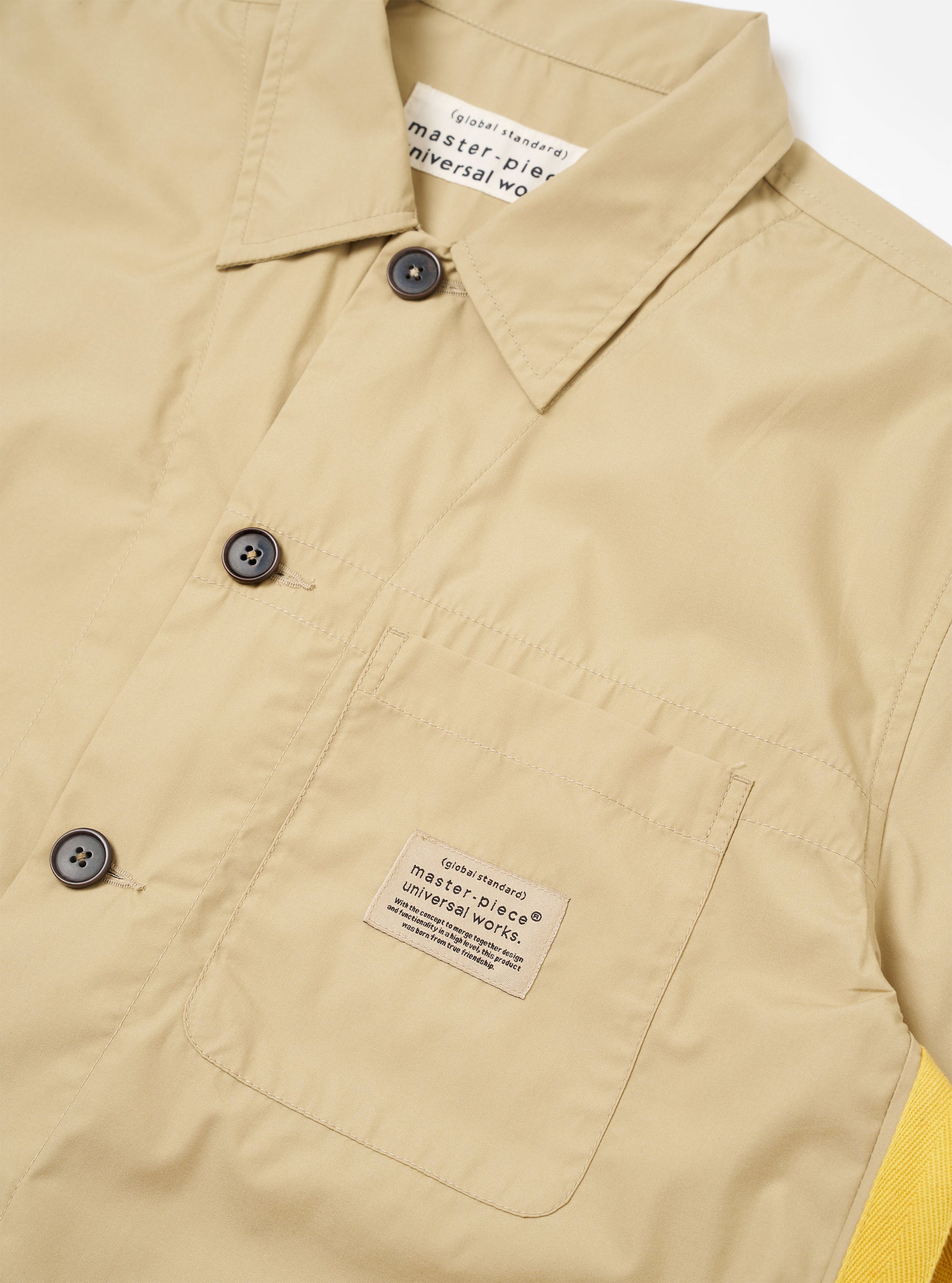 The master-piece x Universal Works Sand Recycled Poly Tech Tech Overshirt is shown from a rear three-quarter angle as a flat lay against a plain white background. The image highlights the back yoke with a box pleat, the short sleeves, and the straight hem with an adjustable elastic drawcord. The sand-colored recycled polytech fabric has a smooth, matte finish.