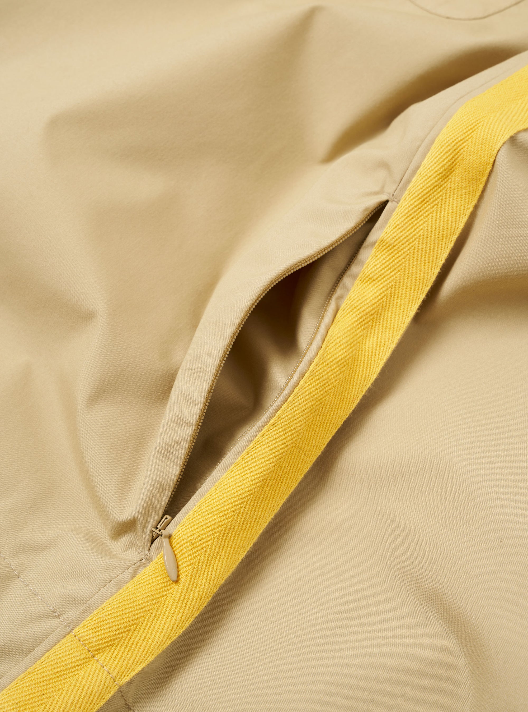 The master-piece x Universal Works Sand Recycled Poly Tech Tech Overshirt is shown in a detail view as a flat lay against a plain white background. The image focuses on the lower part of the garment, highlighting the adjustable hem with its elasticated drawcord and plastic toggle, as well as the clean stitching and the smooth, technical texture of the sand-colored recycled polytech fabric.