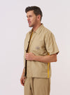 A model wears the master-piece x Universal Works Sand Recycled Poly Tech Tech Overshirt over a light-colored top, standing against a plain white background. The sand-colored overshirt features a structured one-piece collar, a central button-down closure, and a prominent reinforced patch pocket on the left chest. The technical recycled polytech fabric has a smooth, matte finish. Only the upper half of the model is visible in this medium shot.