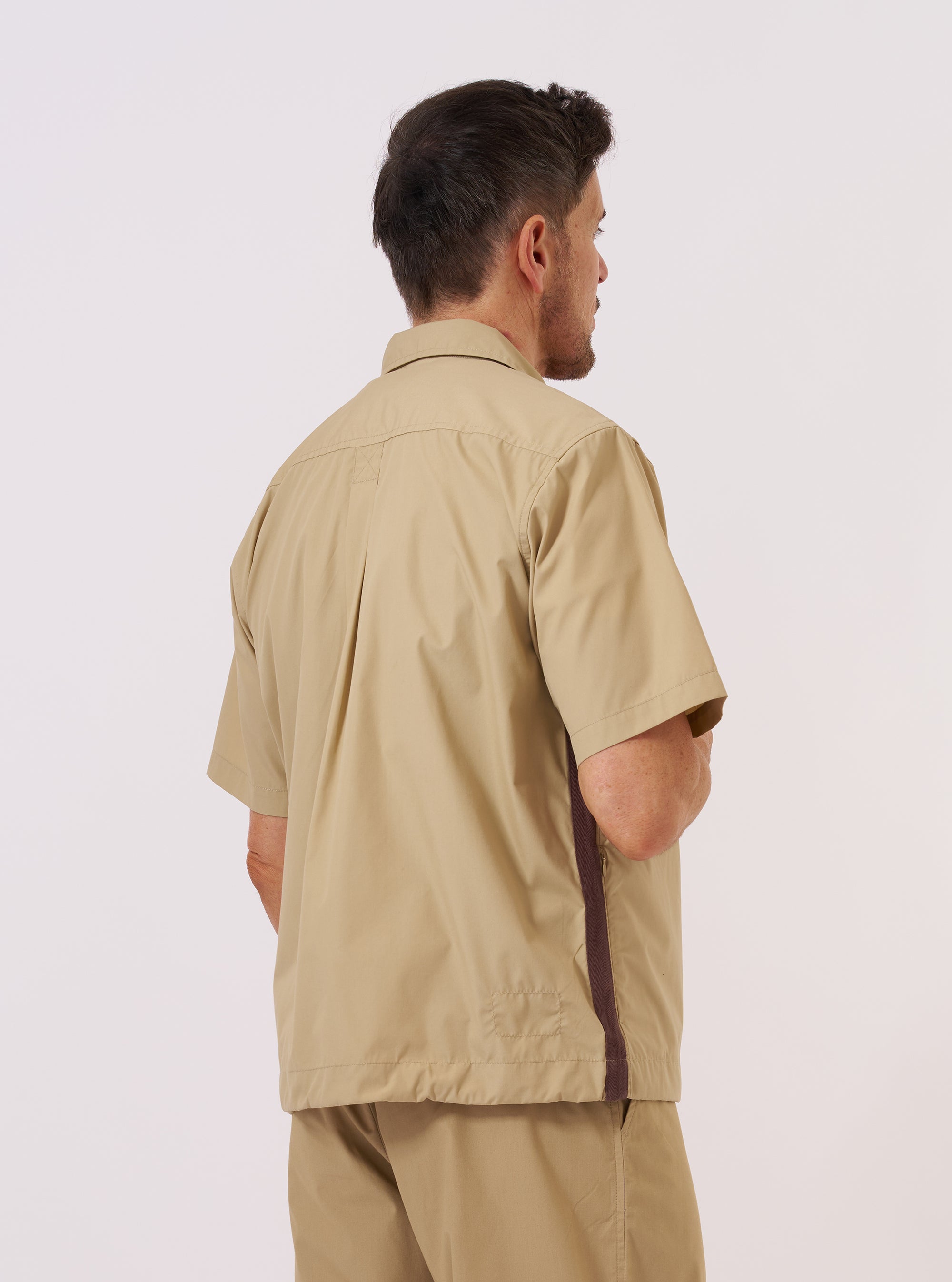 A model is wearing the master-piece x Universal Works Sand Recycled Poly Tech Tech Overshirt, shown from the back against a plain white background. The short-sleeved overshirt features a back yoke with a central box pleat and a straight hem with an adjustable elasticated cord. The garment is a solid sand color with a smooth, technical finish. No mannequin is present in the image.