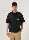 A model is wearing the master-piece x Universal Works Black Recycled Poly Tech Tech Overshirt over a light-colored long-sleeve shirt, shown from a rear three-quarter angle against a plain white background. The overshirt features a clean back yoke with a central box pleat and a straight hem. The black recycled polytech fabric has a smooth, technical finish. No mannequin is present in the image.