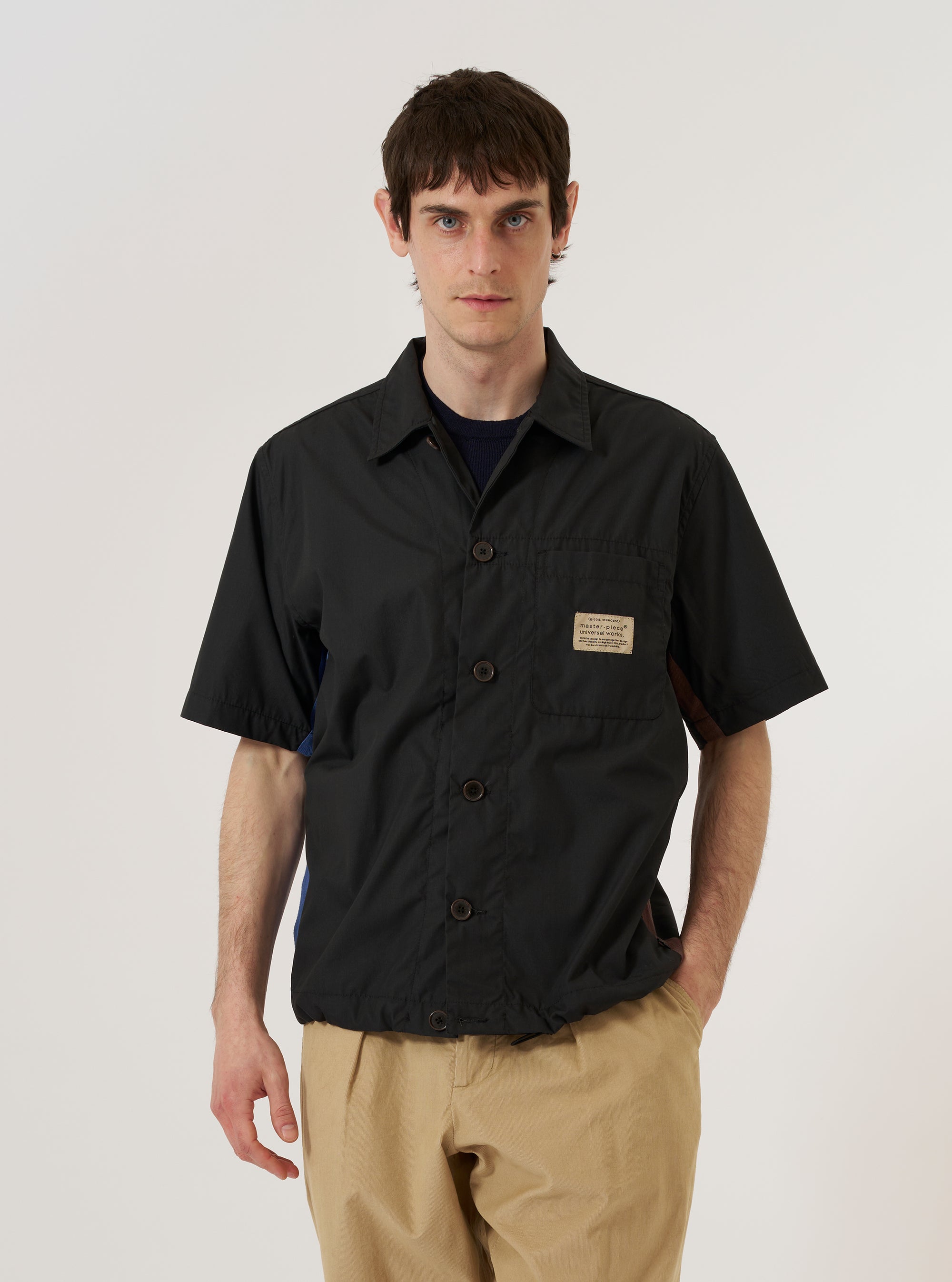 A model is wearing the master-piece x Universal Works Black Recycled Poly Tech Tech Overshirt over a light-colored long-sleeve shirt, shown from a rear three-quarter angle against a plain white background. The overshirt features a clean back yoke with a central box pleat and a straight hem. The black recycled polytech fabric has a smooth, technical finish. No mannequin is present in the image.