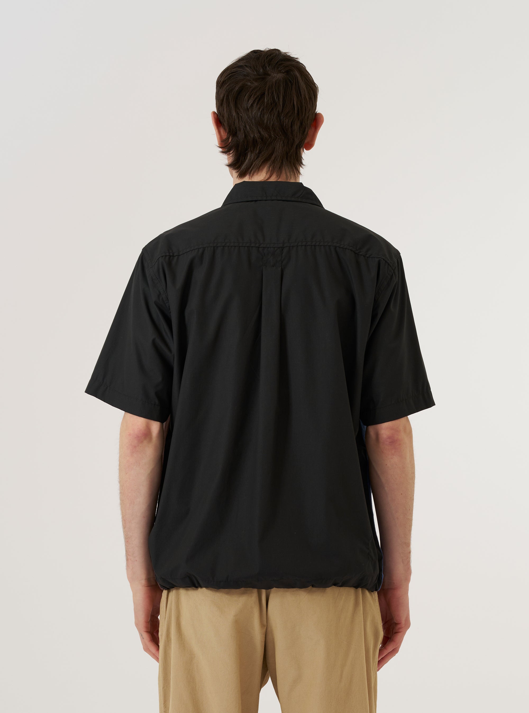 A model wears the master-piece x Universal Works Black Recycled Poly Tech Tech Overshirt over a light-colored top, shown from a rear three-quarter angle against a plain white background. The overshirt features a clean back yoke with a central box pleat and a straight hem. The smooth, matte black technical fabric and the structured one-piece collar are prominent features. No mannequin is present in the image.