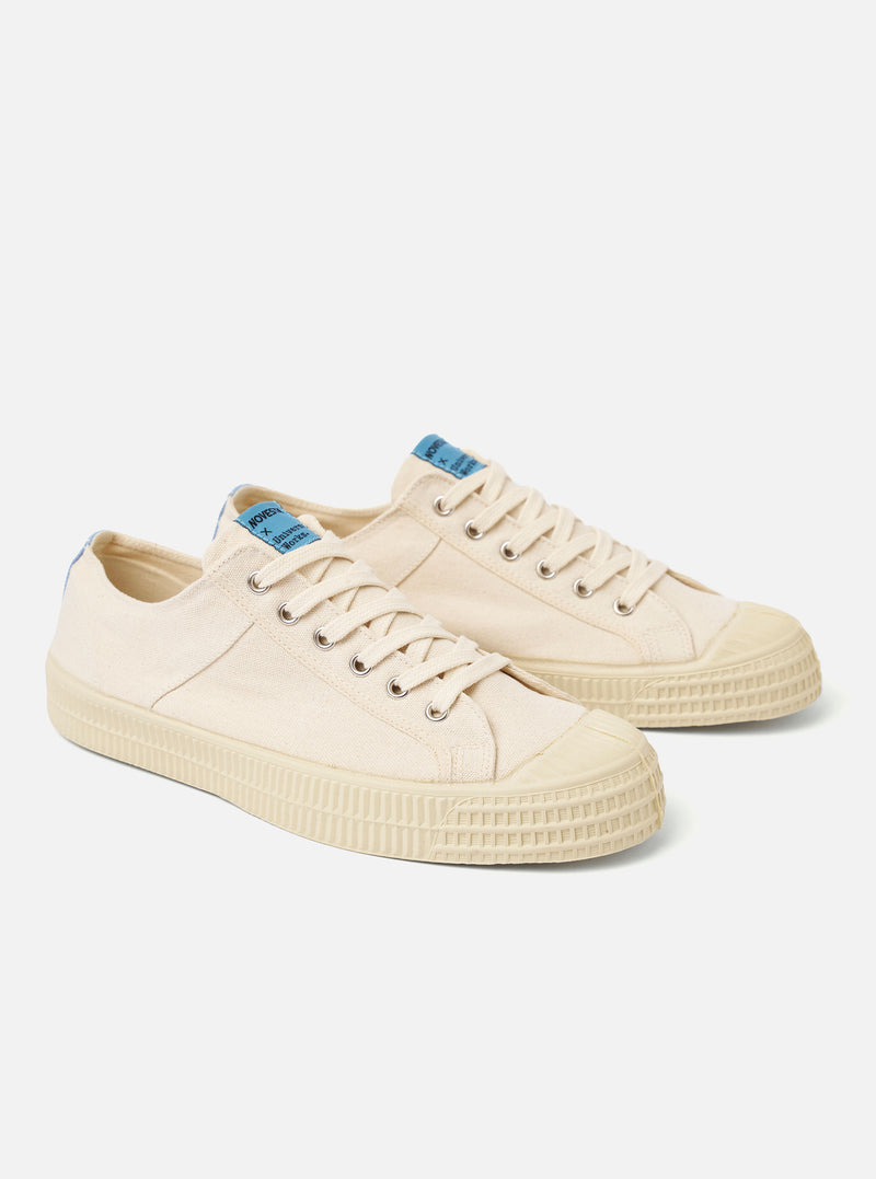 The Novesta for Universal Works Ecru/Pale Blue Canvas Star Master is shown from a side profile view against a white background. The low-top sneaker features an ecru cotton canvas upper with contrast stitching, metallic eyelets, and a signature chunky vulcanized rubber sole in ecru. A pale blue pull tab is visible on the heel, and a dual-branded woven tag is attached to the tongue.