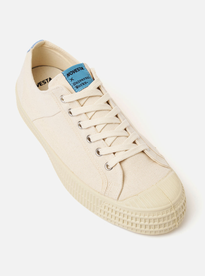 The Novesta for Universal Works Ecru/Pale Blue Canvas Star Master is shown from a side profile against a white background. The sneaker features an ecru cotton canvas upper with tonal contrast stitching and chrome metallic eyelets. A prominent pale blue fabric strip is attached to the heel. The shoe is constructed with a chunky, textured ecru vulcanized rubber sole and a matching rubber shell toe.
