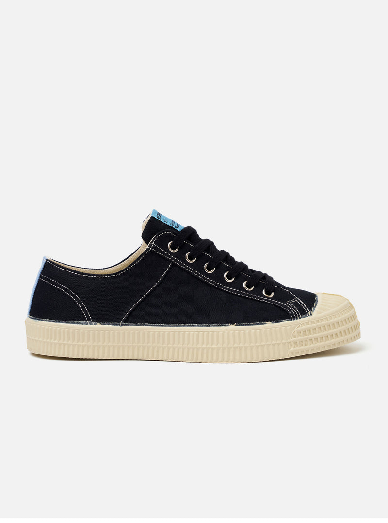 The Novesta for Universal Works Black/Pale Blue Canvas Star Master is shown from a side profile as a flat lay against a white background. The low-top sneaker features a black cotton canvas upper with contrast white stitching and metallic eyelets. A signature chunky ecru vulcanized rubber sole and toe cap are prominent, with a pale blue flash detail on the heel.