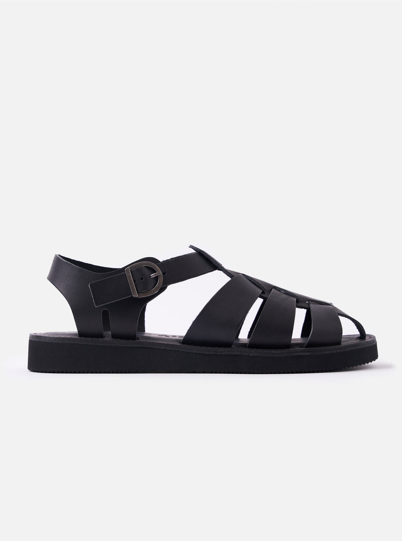 The Organic Handloom Black Leather Gurkha is shown as a flat lay against a white background. The footwear features a braided upper design with multiple black leather straps and a silver-tone buckle closure on the side. The shoe is finished with a chunky, black foam rubber outsole.