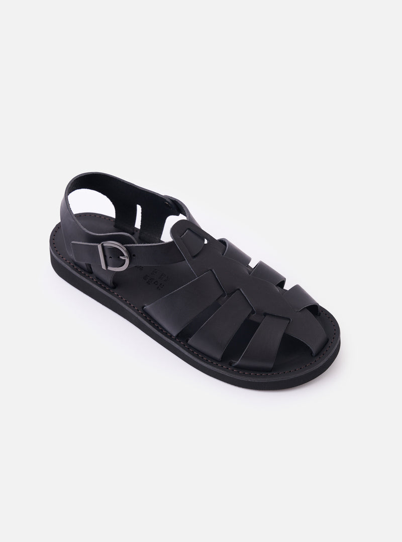 The Organic Handloom Black Leather Gurkha is shown from a side profile as a flat lay against a plain white background. The sandal features a matte black leather upper with a traditional woven strap design, a closed toe, and an adjustable ankle strap with a silver-tone buckle. The thick, black foam rubber outsole is prominent, showing its textured edge.
