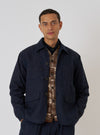 A model poses in an Indigo Brushed Denim Labour Jacket against a white backdrop. The jacket is paired with a patterned vest and dark shirt, showcasing its versatile fit.