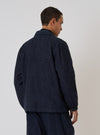 A man poses in the Indigo Brushed Denim Labour Jacket against a plain background. The jacket is a dark indigo color and appears to be made of denim. The fit is relaxed.