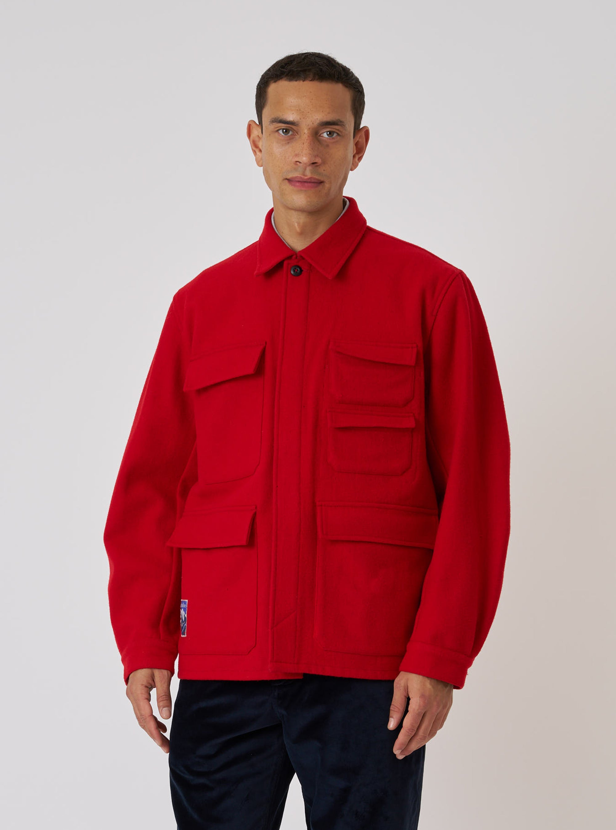 A model poses in front of a white backdrop, wearing the Embroidered Red Mowbray Military Fatigue Jacket. The jacket is a vibrant red with multiple pockets.