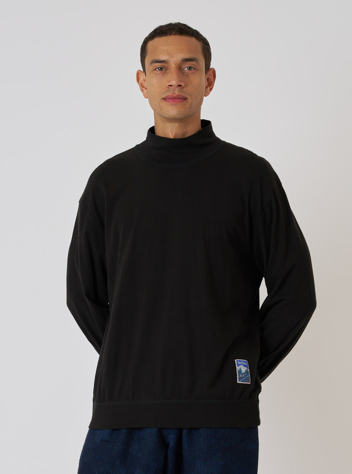Studio shot on white of a model wearing a Black Single Jersey Funnel Neck Tee. The tee is black and has a funnel neck. The model is fair-skinned and is wearing blue pants.