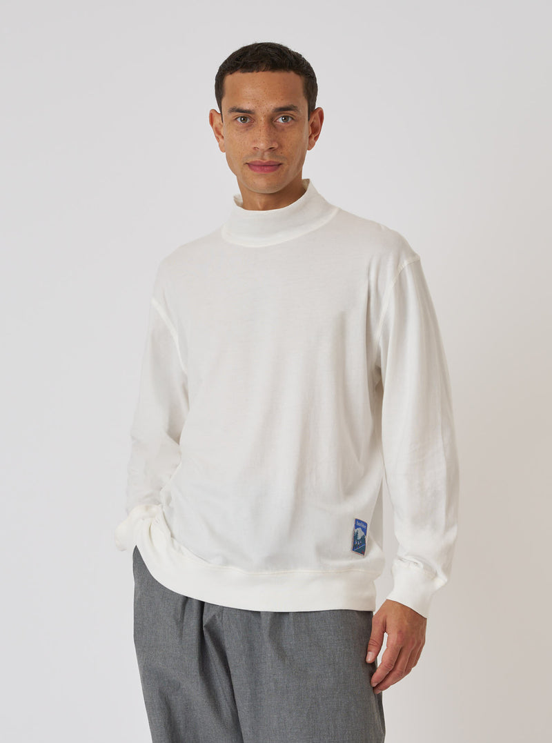 A model poses in an Ecru Single Jersey Funnel Neck Tee against a white backdrop. The tee is paired with grey trousers. The fit is relaxed and casual.