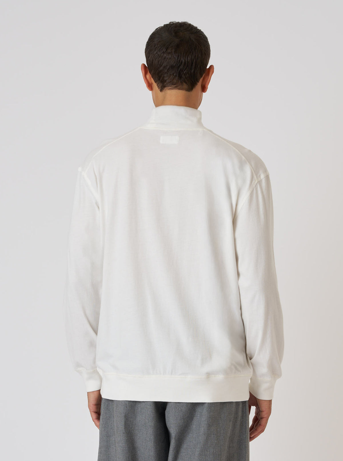 Back view of a model wearing an Ecru Single Jersey Funnel Neck Tee. The tee is a light cream color and has long sleeves. The background is a plain white studio backdrop.