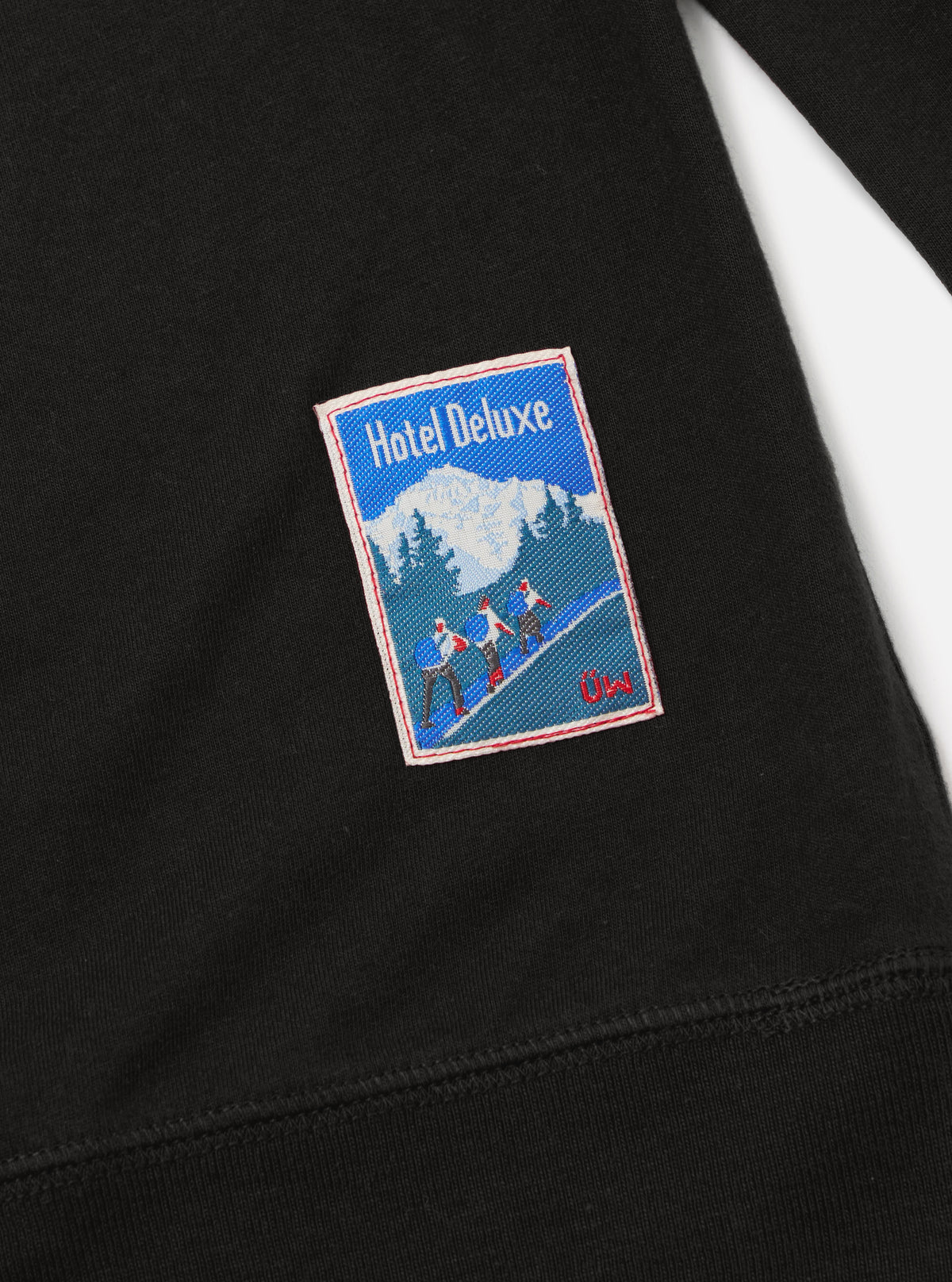 Close up of the black Single Jersey Funnel Neck Tee showing the fabric and a Hotel Deluxe patch on a white background.
